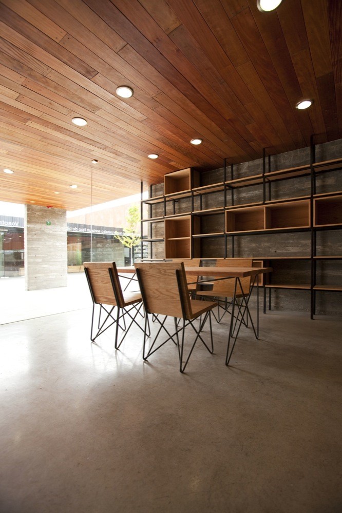 Dining table with wire-legged wooden chairs before a steel-framed shelving unit, under a timber plank ceiling with recessed lights