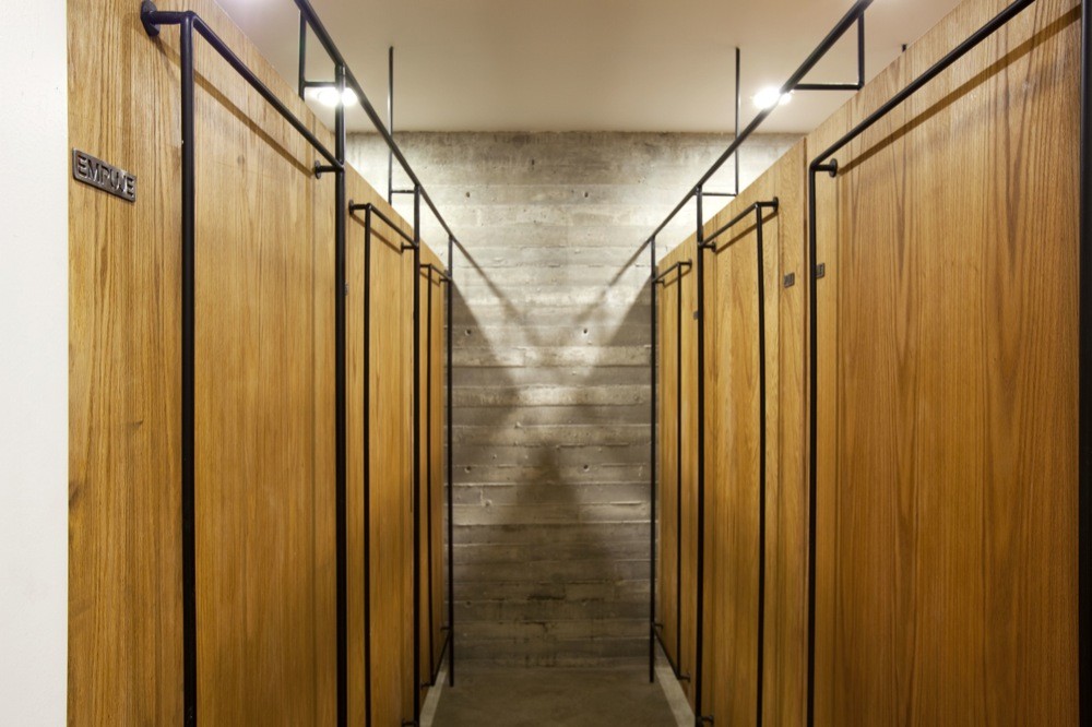 Narrow corridor lined with oak changing-room doors and black steel rails leading to a board-formed concrete end wall