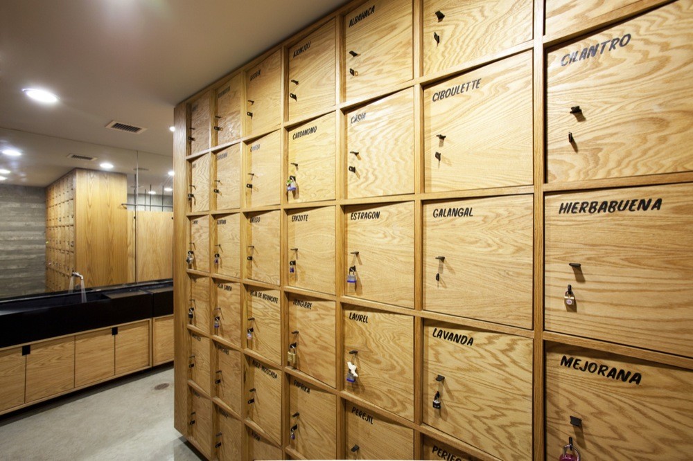 Wall of oak lockers hand-labelled with herb names such as cilantro, laurel and lavanda, beside a black basin counter
