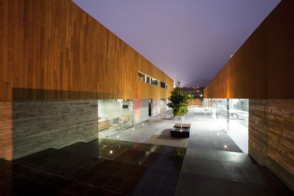 Courtyard between timber-clad volumes on board-formed concrete bases, with a lone tree in a planter under a dusk sky