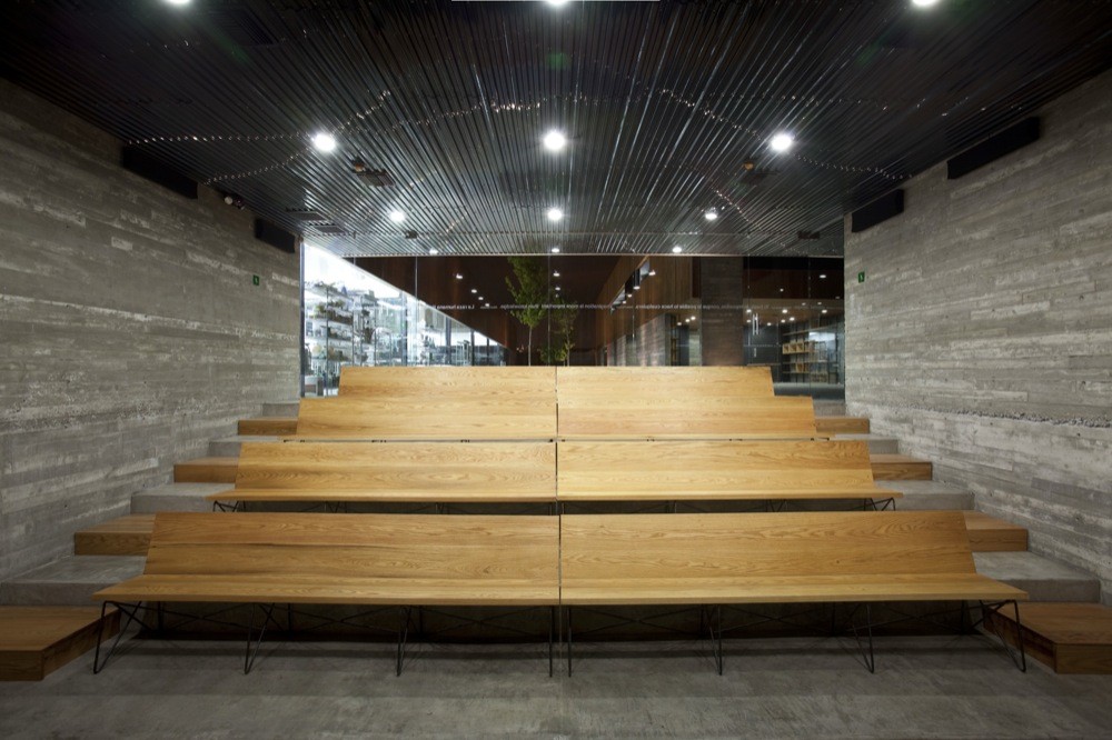 Tiered timber bench seating between board-formed concrete walls, beneath a dark slatted ceiling with recessed ceiling lights