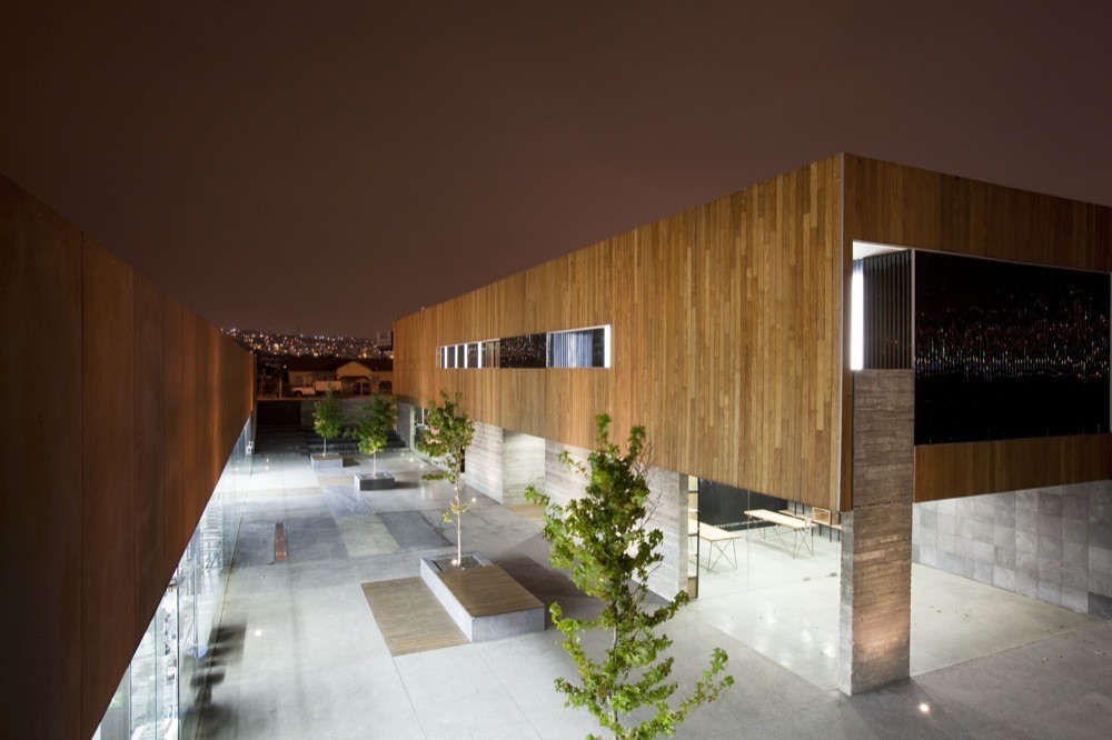 Raised view of the night courtyard with planted trees, timber-clad upper volume and a board-formed concrete column