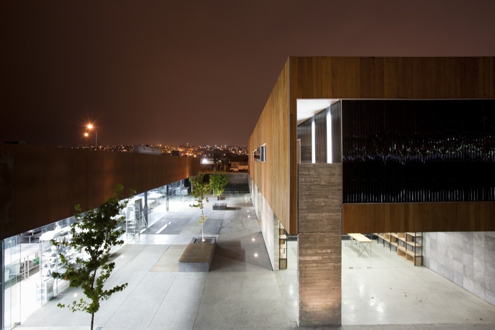 Night view across the paved courtyard toward a concrete column and timber box, with distant city lights on the horizon