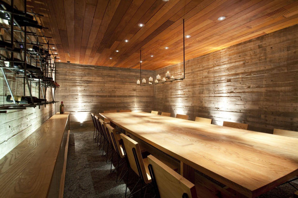 Long timber dining table with chairs in a concrete-walled room, wine rack on the left and candle chandelier overhead
