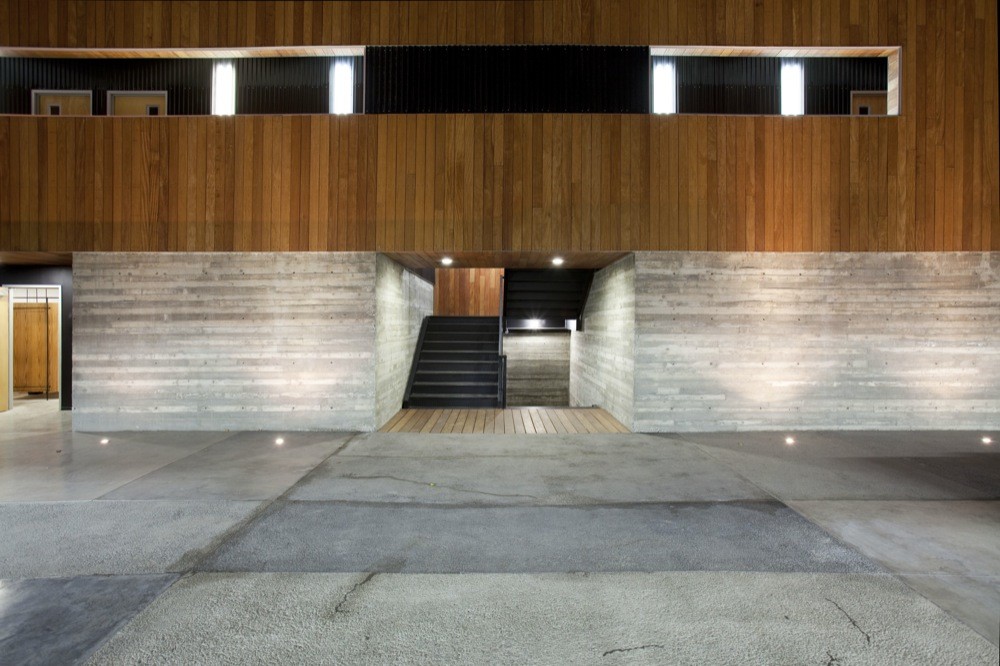 Timber-clad upper floor above board-formed concrete walls, with a dark steel stair rising from a pale paved forecourt at night
