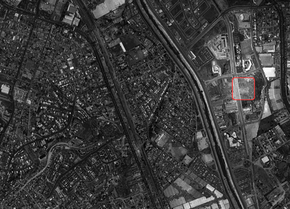 Black and white aerial site plan photograph with a red outline marking an empty lot beside a highway and dense neighborhood blocks