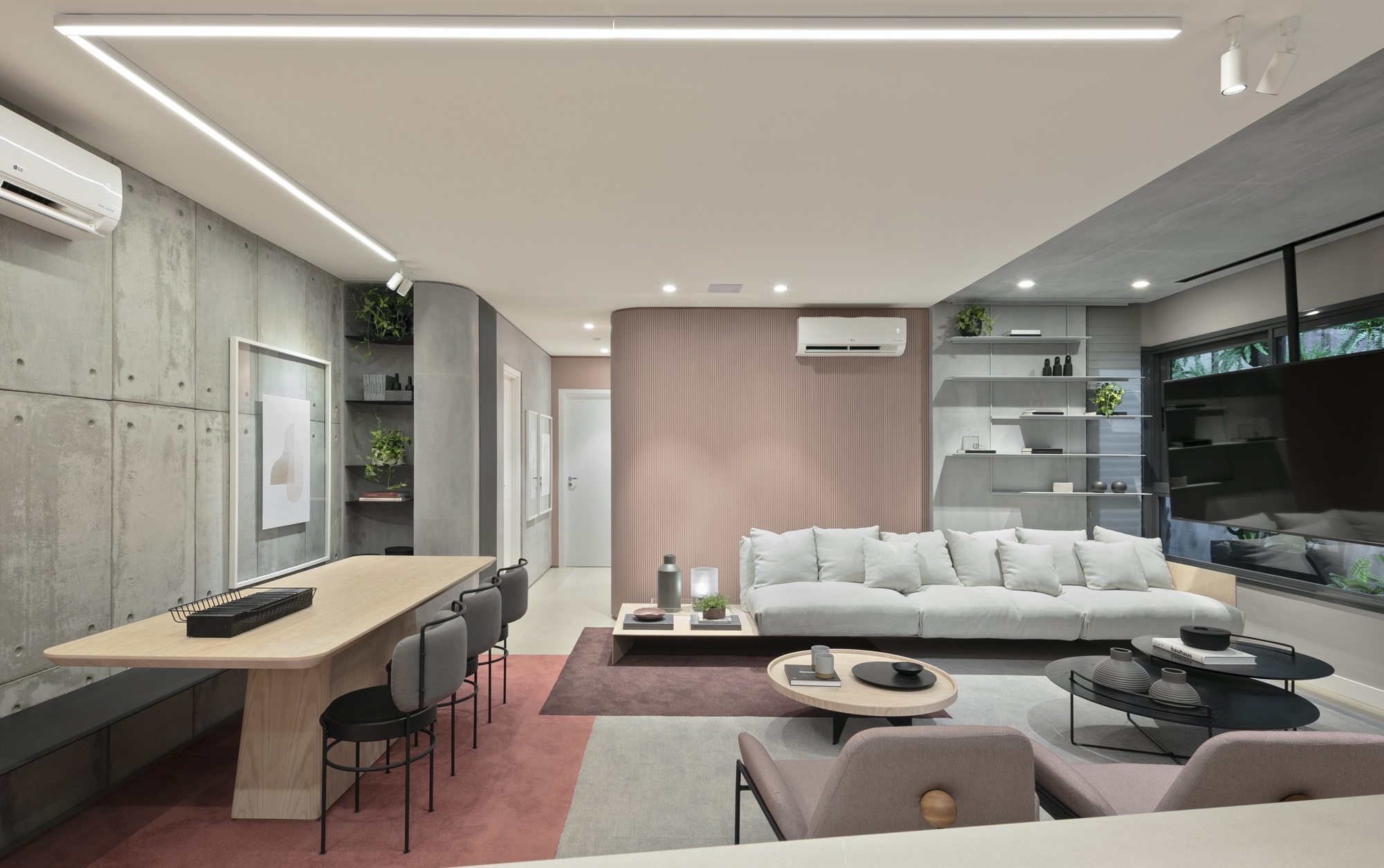 Living area with board-marked concrete wall, long timber table and grey sofa against a blush pink panel, lit by a linear ceiling fixture