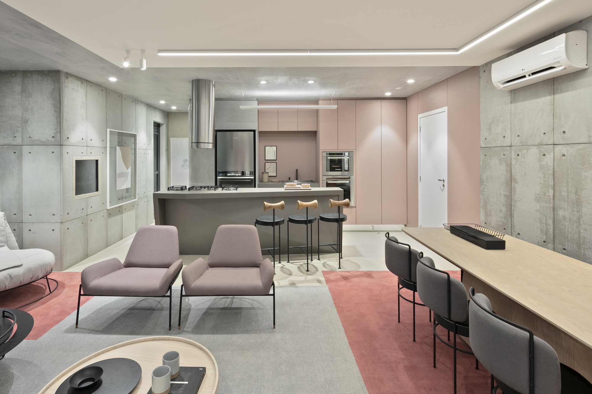 Open kitchen with a concrete island, wood-topped stools and pink cabinetry, seen past two mauve armchairs on a grey rug