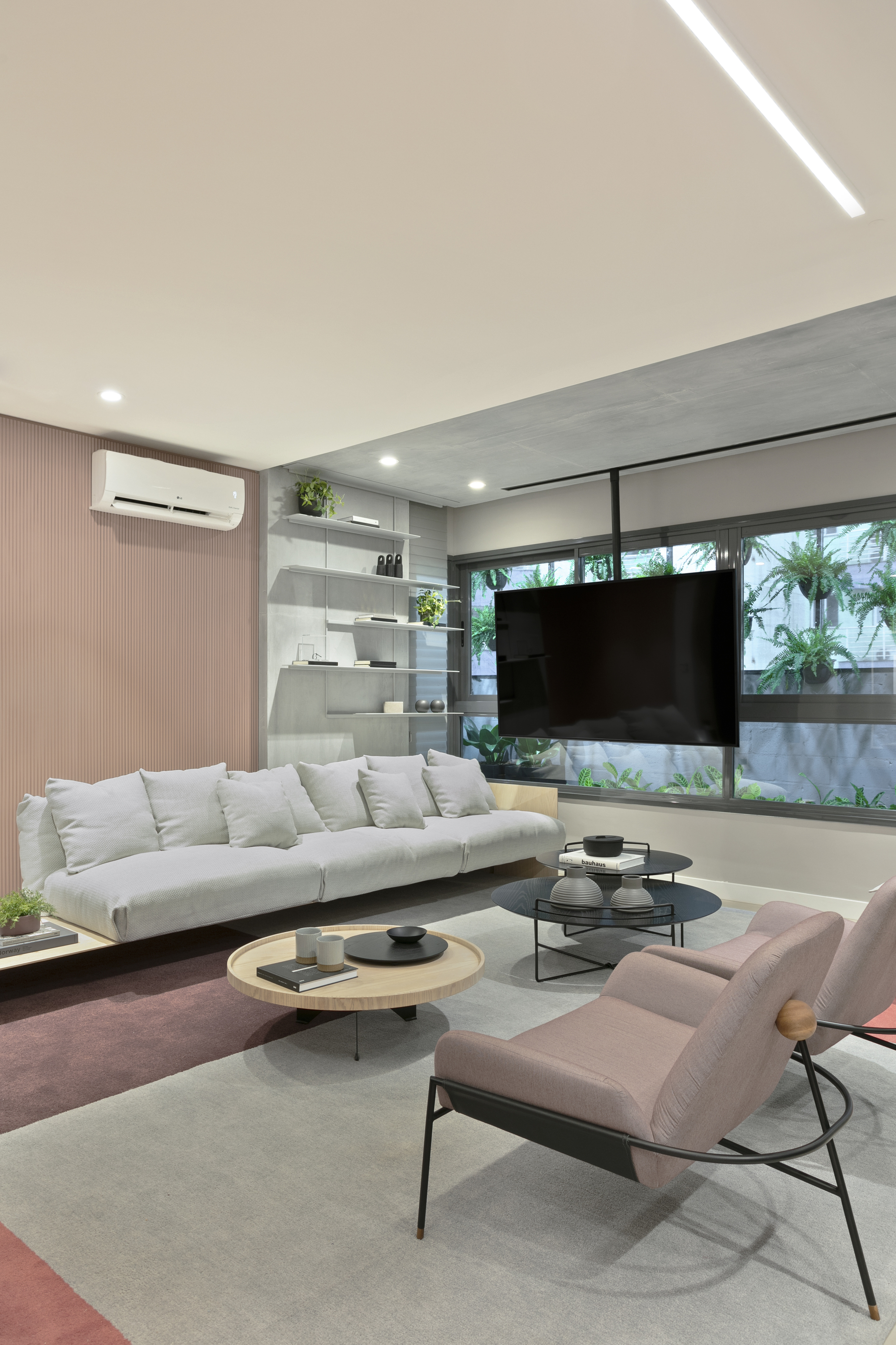 Pink lounge chair and round coffee tables facing a wall-mounted television set against a window of green plants