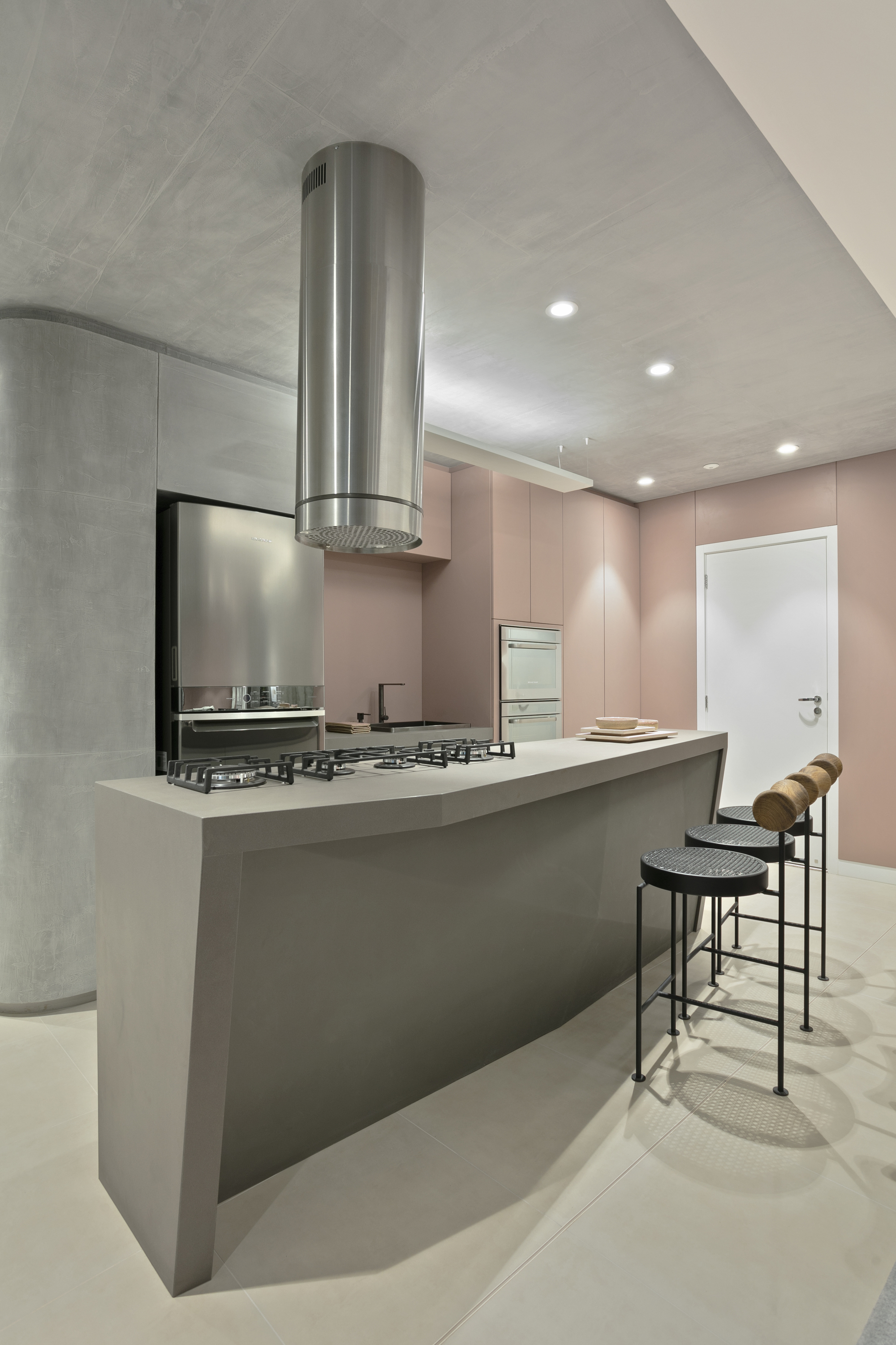 Angular concrete kitchen island with gas hob, steel cylindrical hood and black stools beside pink cabinetry