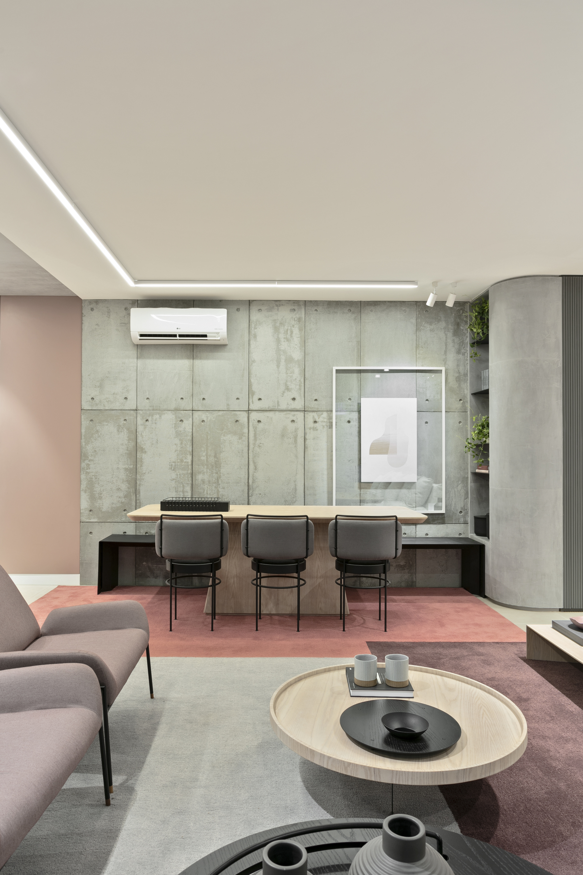 Living area with a board-formed concrete panel wall, grey upholstered dining chairs and a coral rug under a recessed linear ceiling light