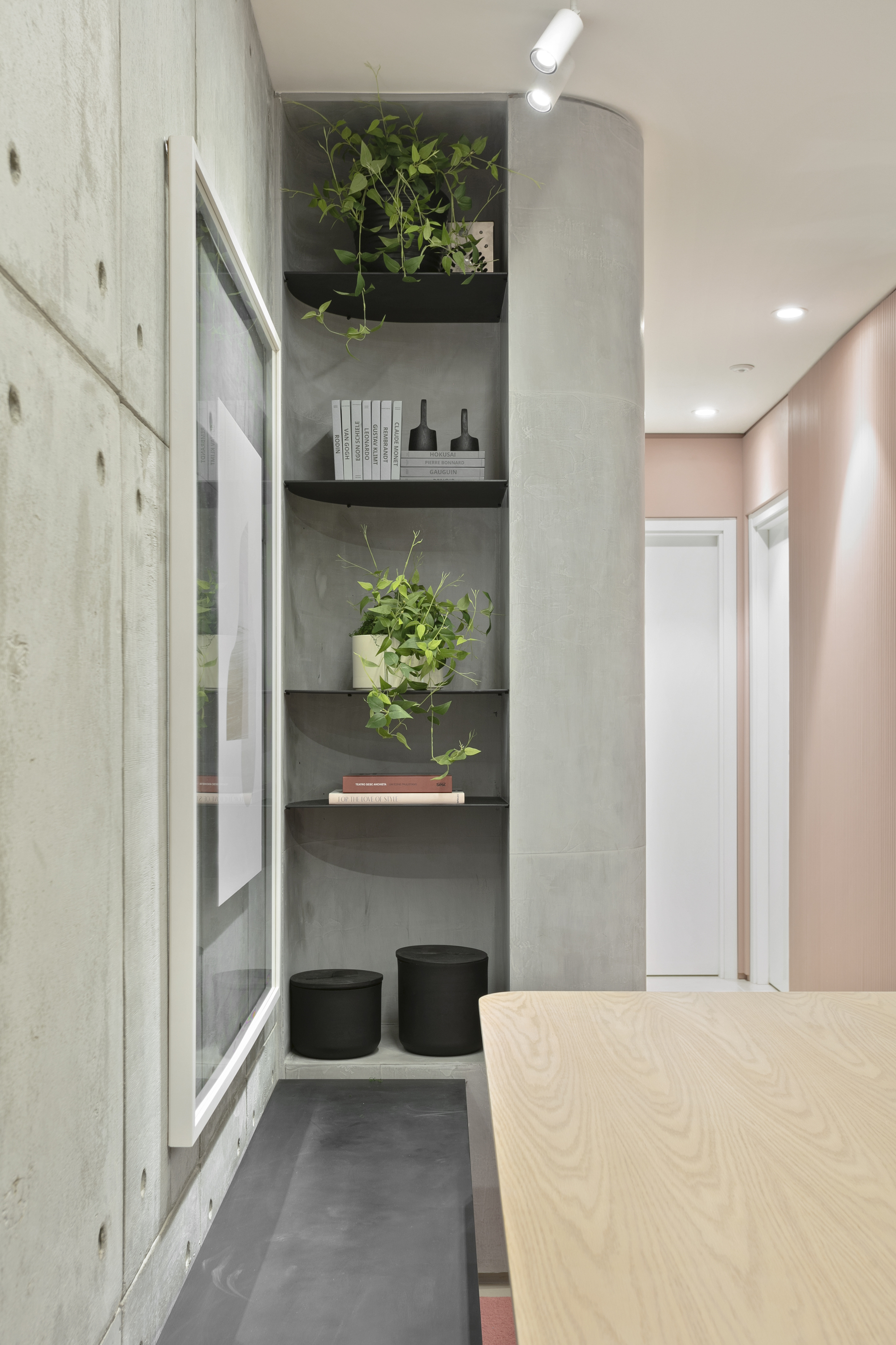 Curved concrete column with black metal shelves holding trailing plants, books and two black stools beside a pale wood tabletop