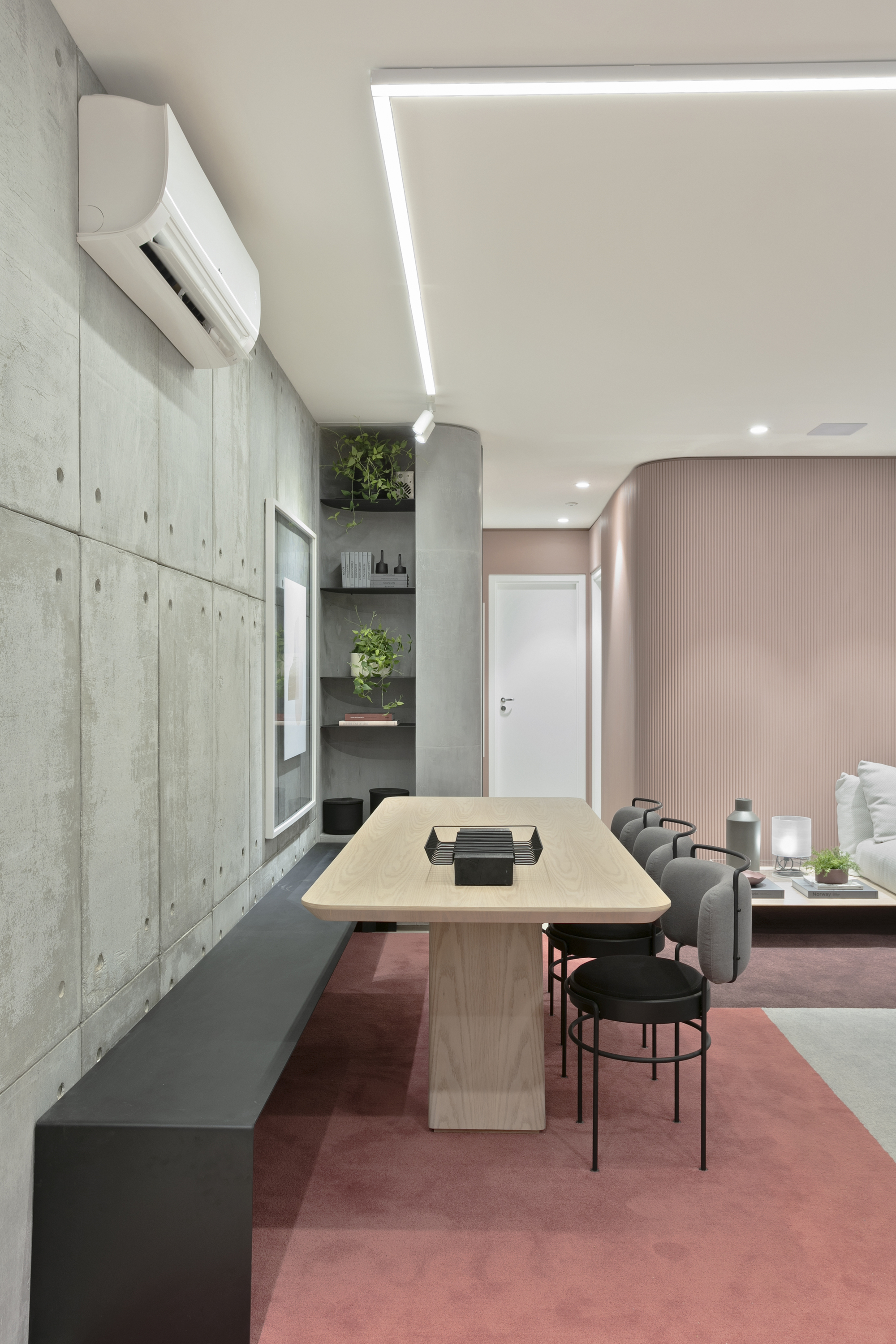 Long light wood dining table with black chairs and a dark bench against the concrete panel wall, with a fluted pink wall beyond