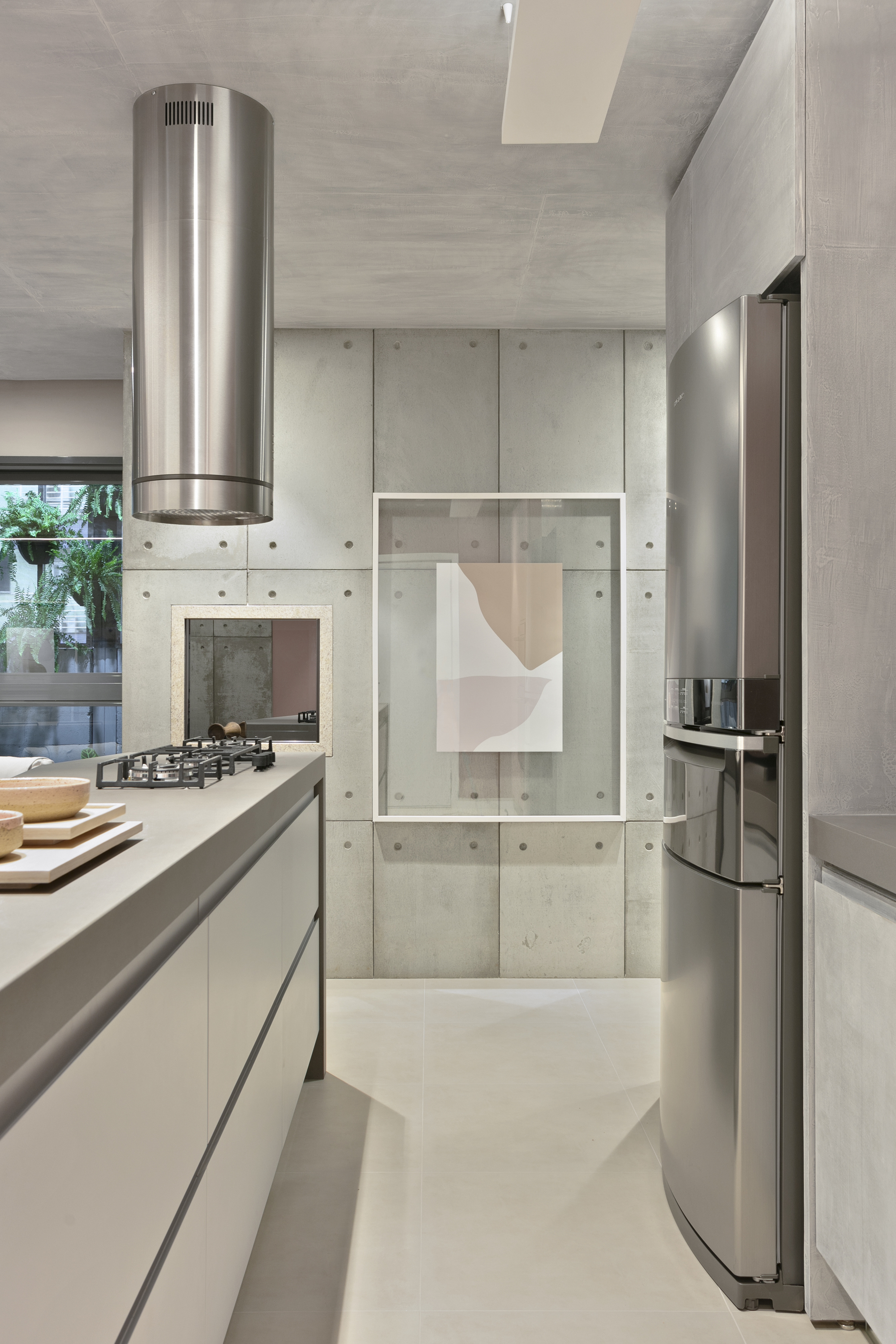 Kitchen corridor with a cylindrical steel hood, stainless refrigerator and a framed print hung over a concrete panel wall with a leafy window at left