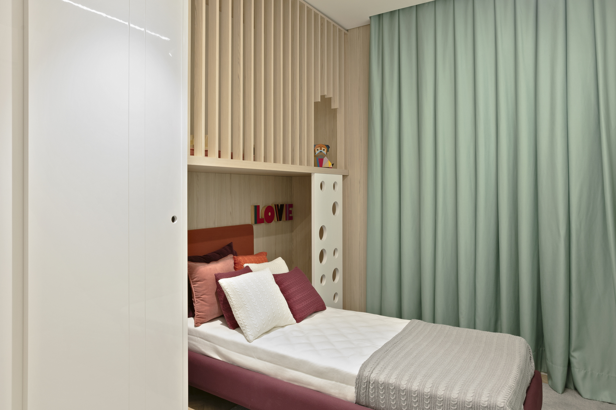 Child's bed tucked under a slatted timber loft with a perforated white panel, beside a long sage green curtain