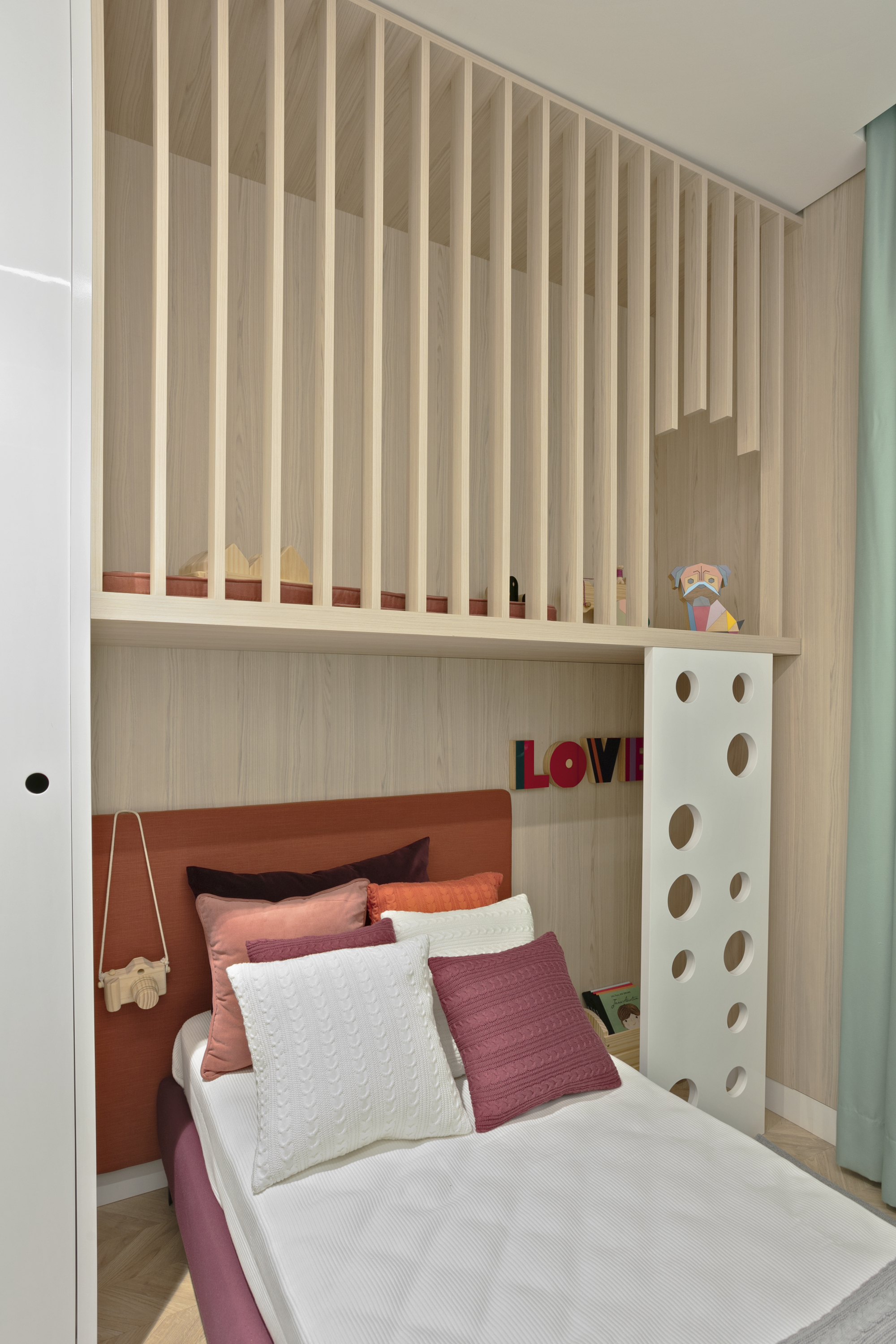 Vertical view of the slatted wood loft above a child's bed, with stacked cushions, a camera-shaped bag and a LOVE sign