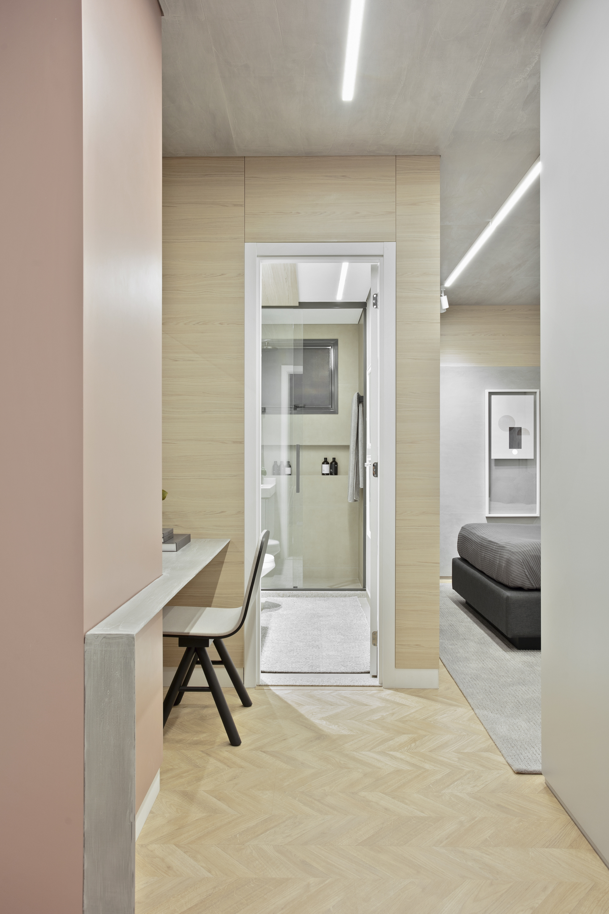Hallway with a built-in desk and chair leading past a glass-door bathroom, with a bedroom glimpsed beyond under linear ceiling lights