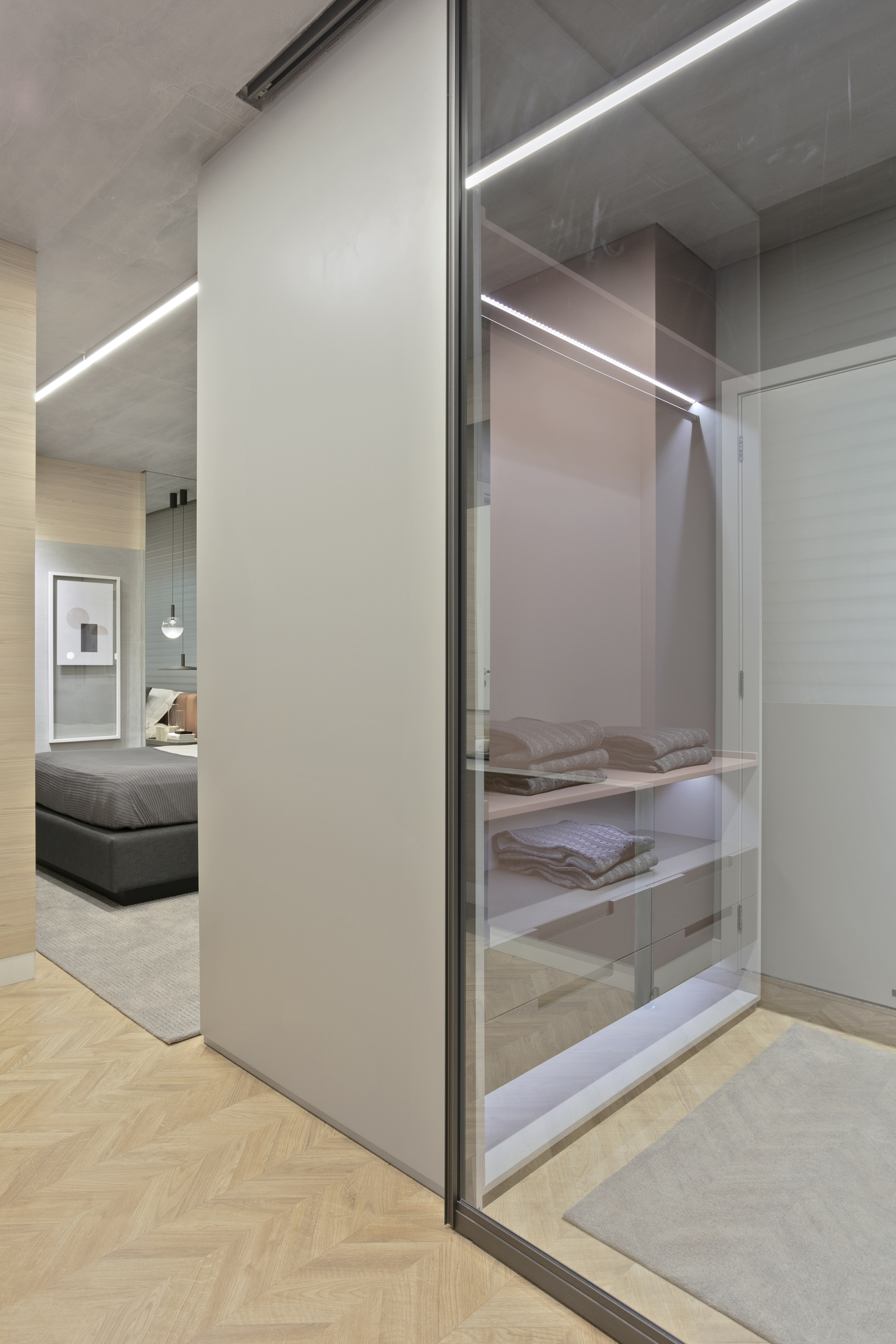 Glass-fronted walk-in closet with lit shelves of folded towels, seen past a sliding panel toward a bedroom with herringbone flooring