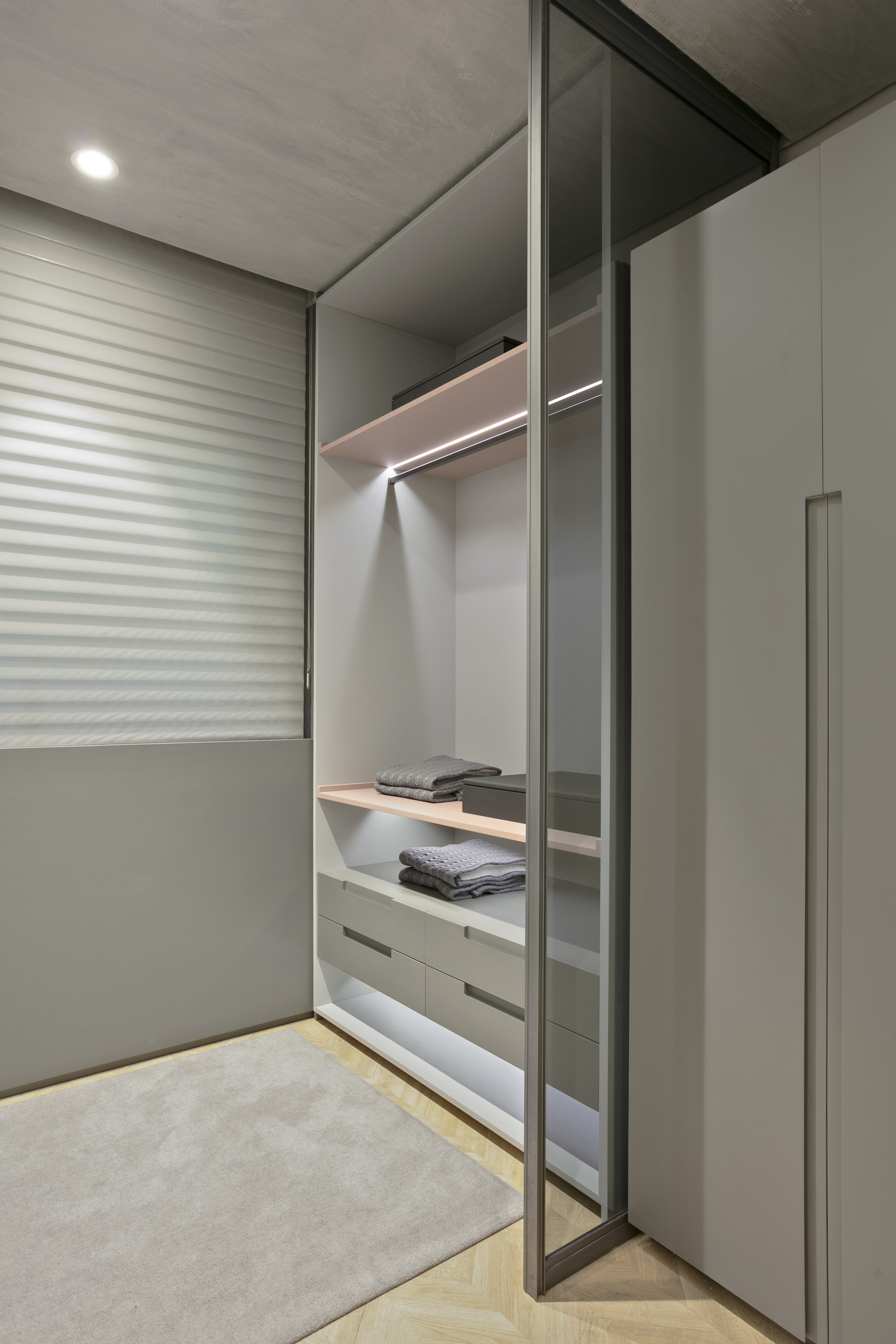 Walk-in closet with glass door, lit hanging rail, shelves and drawers, beside a roller shutter and a pale grey rug