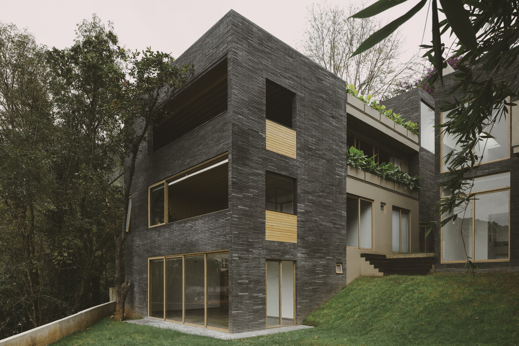 Corner of the dark brick volume with timber-framed windows and planted terraces, set on a grassy slope under overcast sky