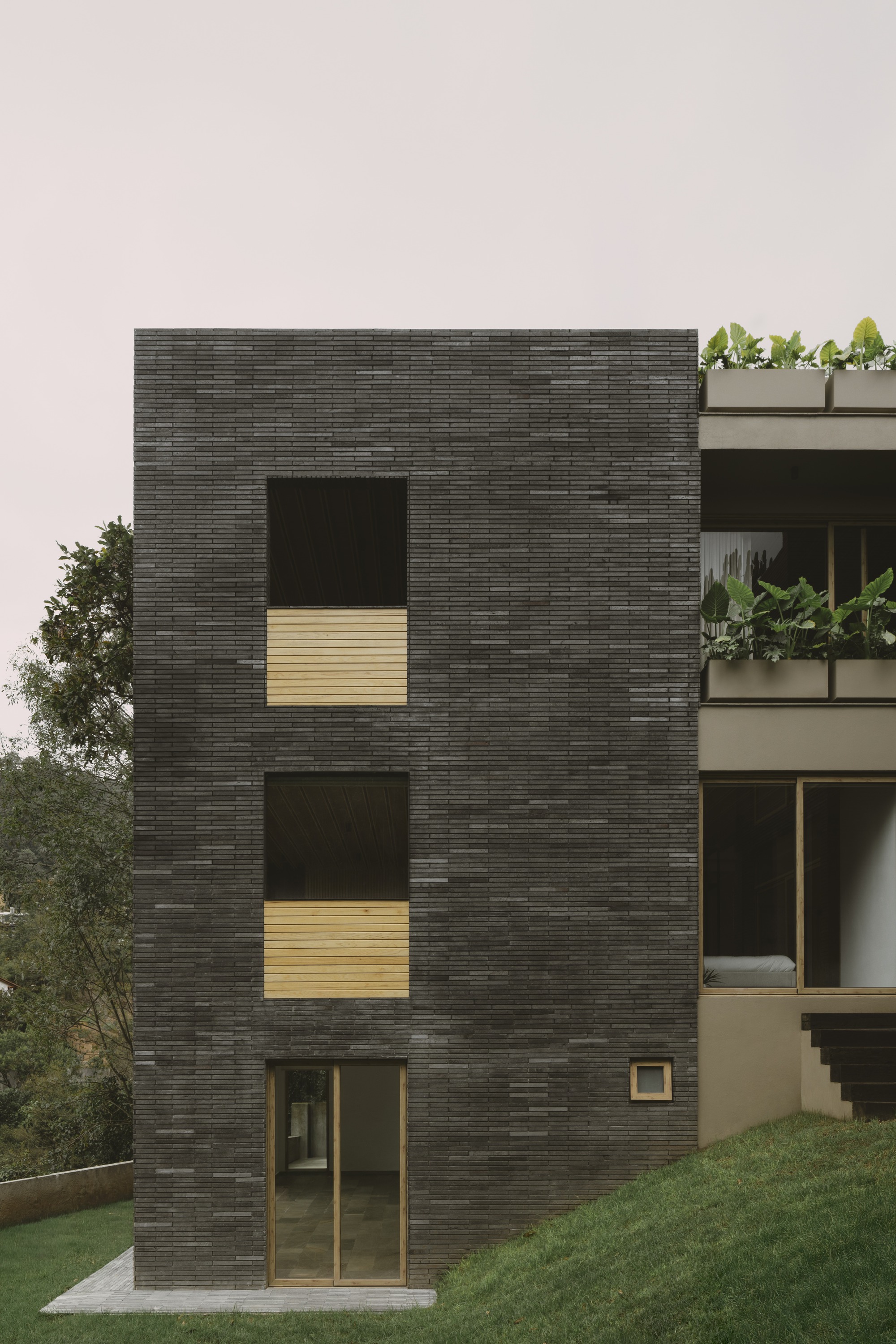 Dark brick tower elevation with two recessed openings, timber infill panels and a small square window beside a grassy bank