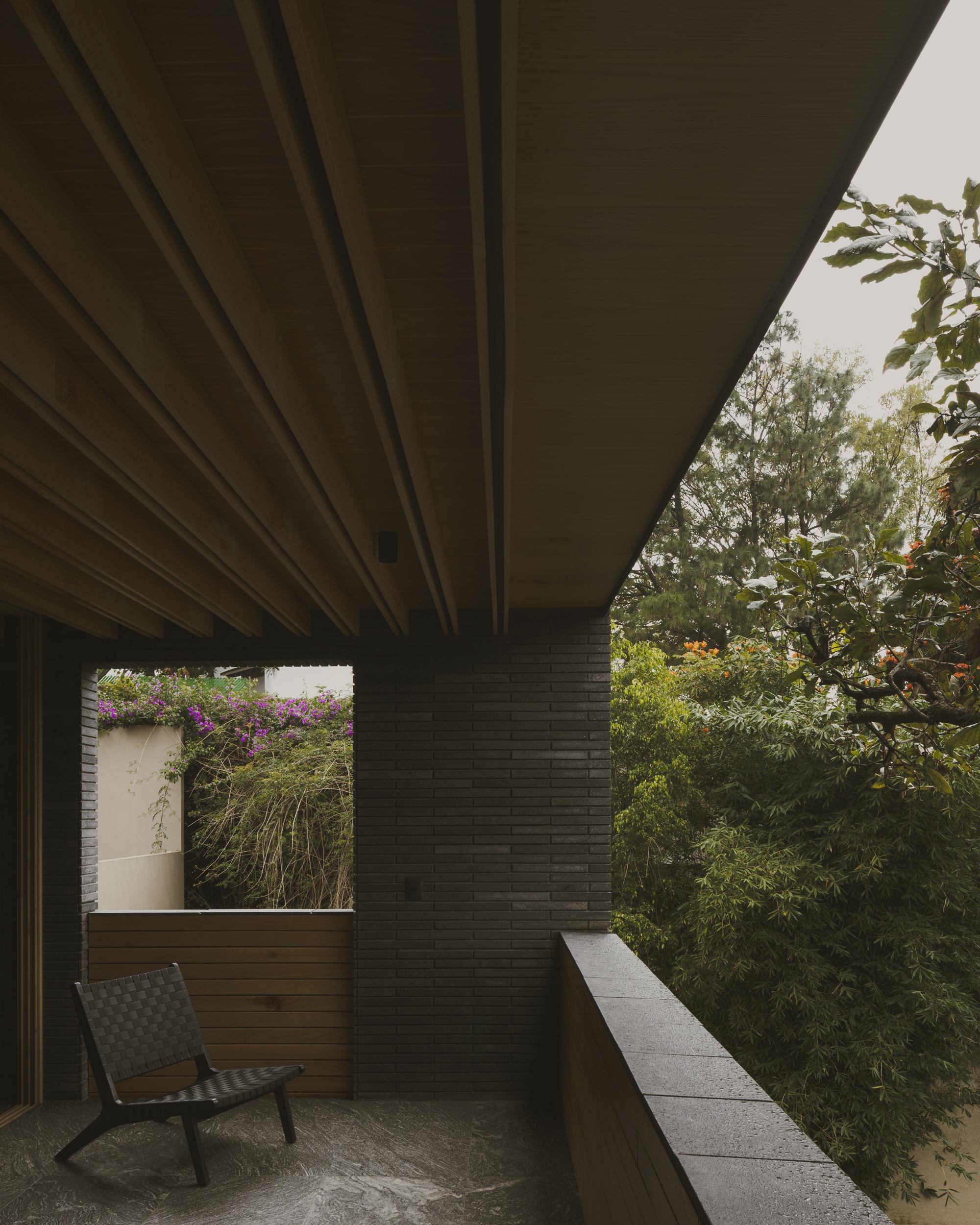 Covered terrace with timber ceiling beams, a dark brick wall, a woven chair and flowering vines glimpsed beyond in grey light