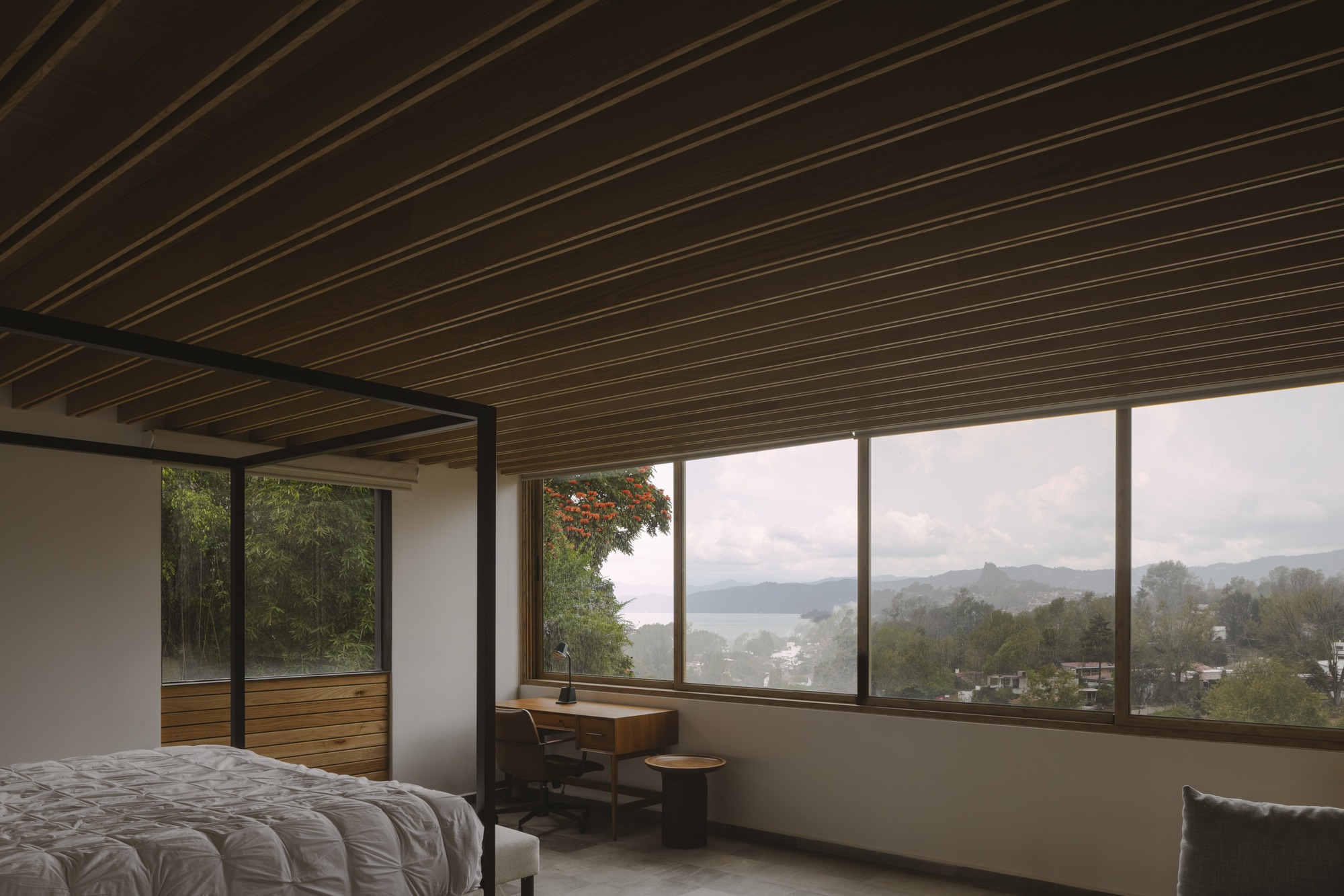 Bedroom with a four-poster bed, timber ceiling beams and a corner window framing a distant lake and hills under cloud