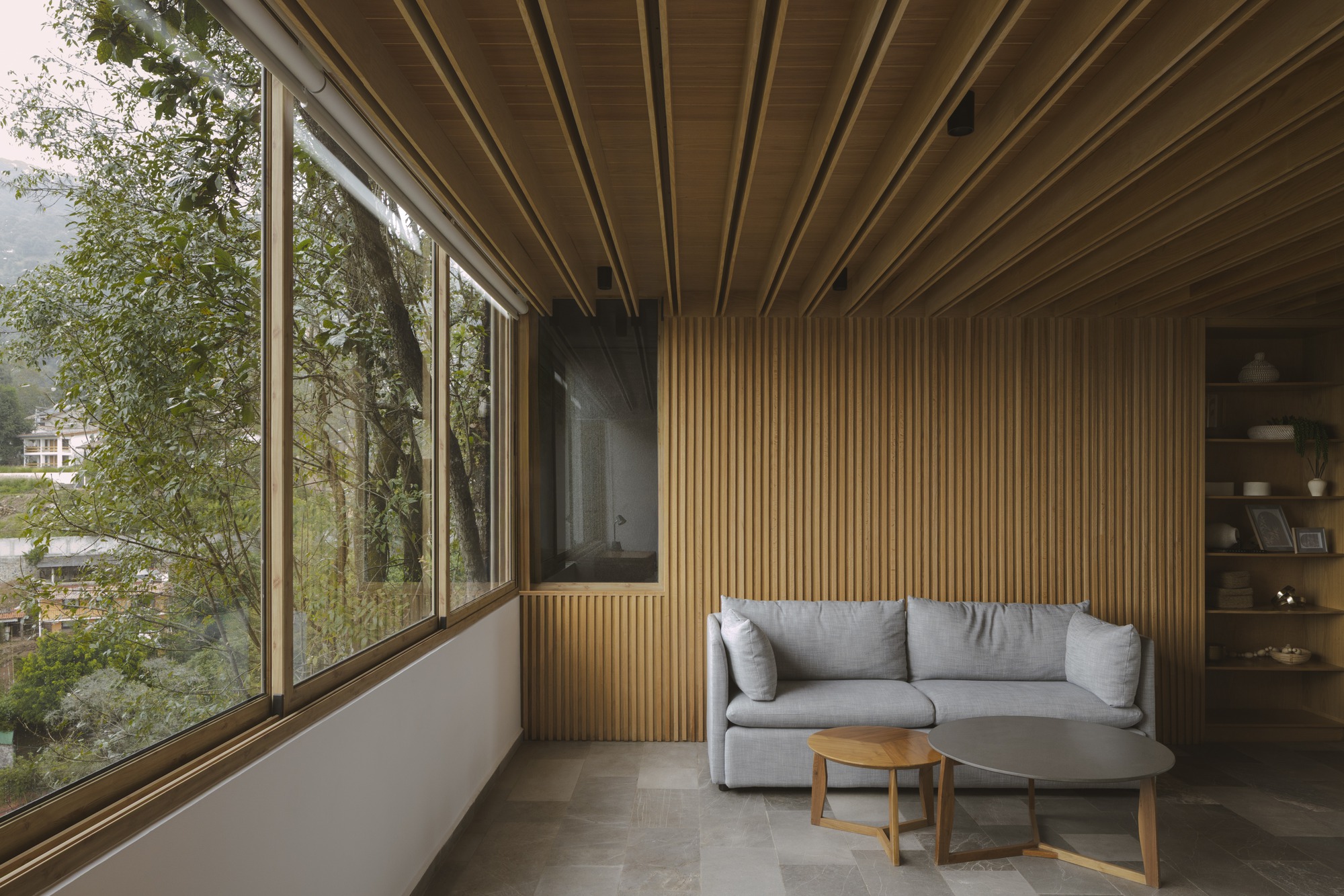 Sitting area with a grey sofa against a vertical timber slat wall, beside a tall window onto trees
