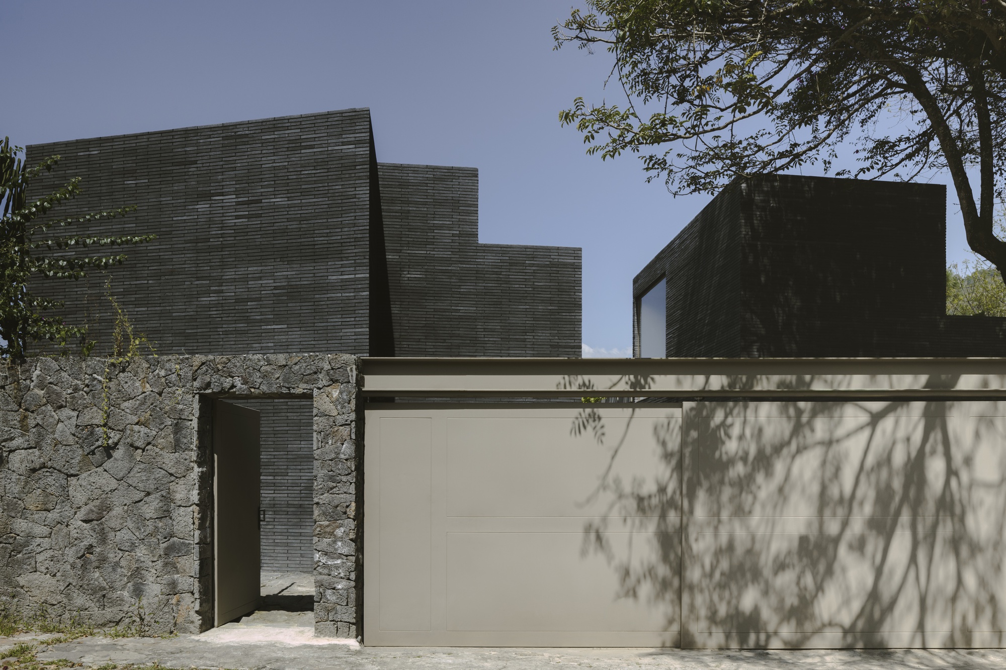 Street facade of dark brick volumes behind a rough volcanic stone wall and taupe gate, tree shadows falling across the panels