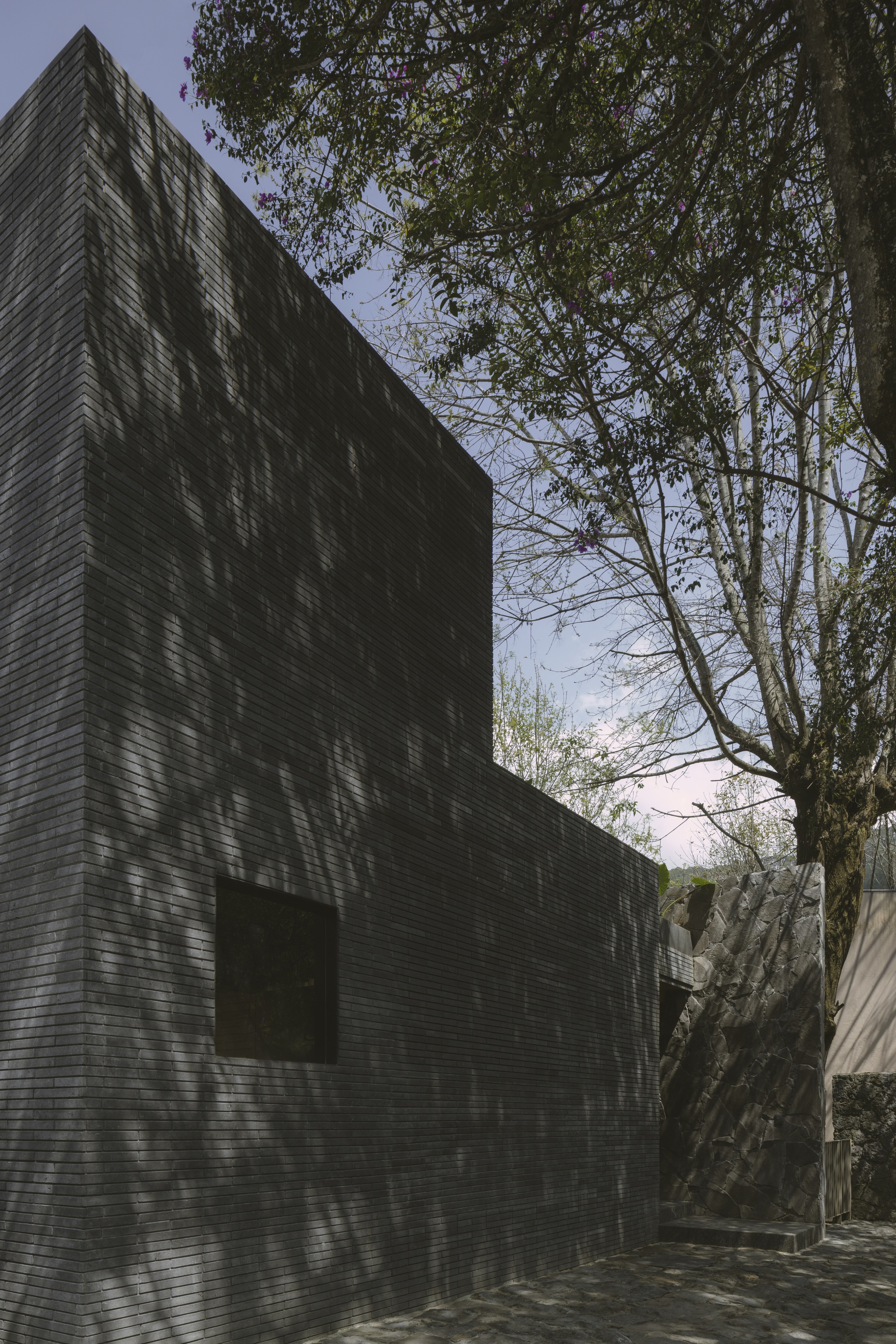 Side elevation of the dark brick tower with a square window, dappled by shadows of a flowering tree