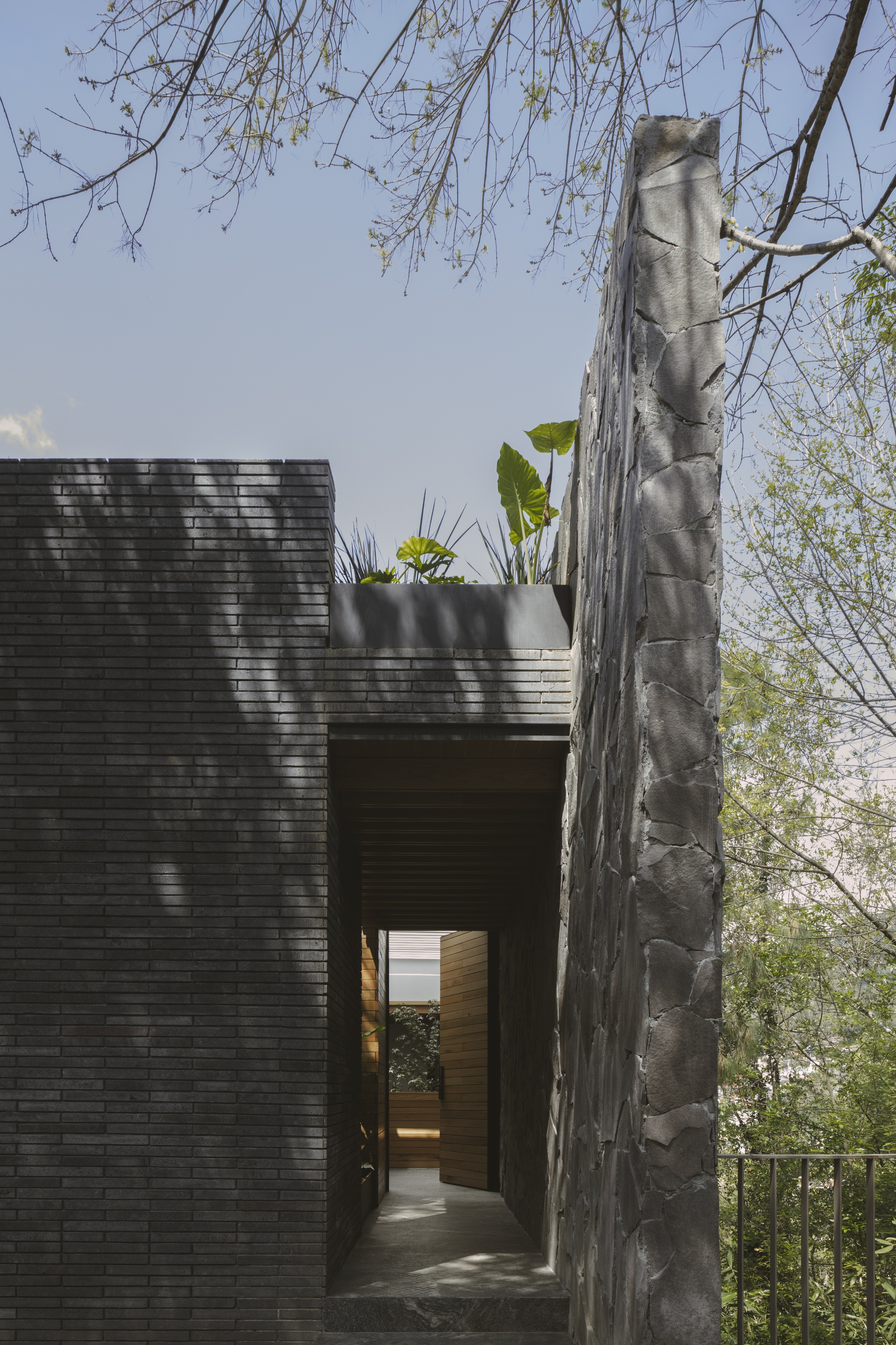 Narrow entry passage between a dark brick wall and a tall stone wall, with plants on the roof above