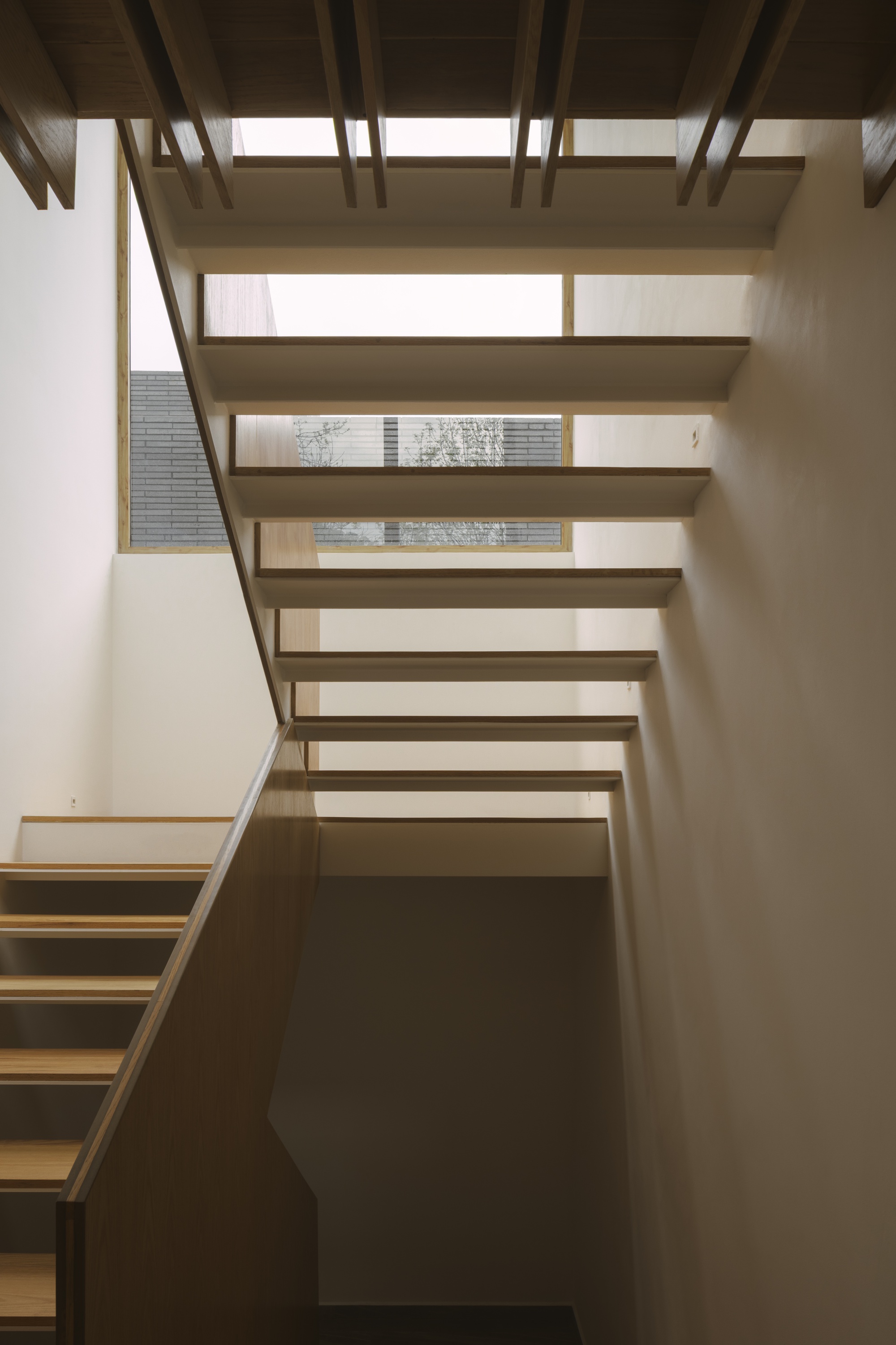 Timber stair with floating treads rising beside a pale wall and a window onto neighboring brick, in soft daylight