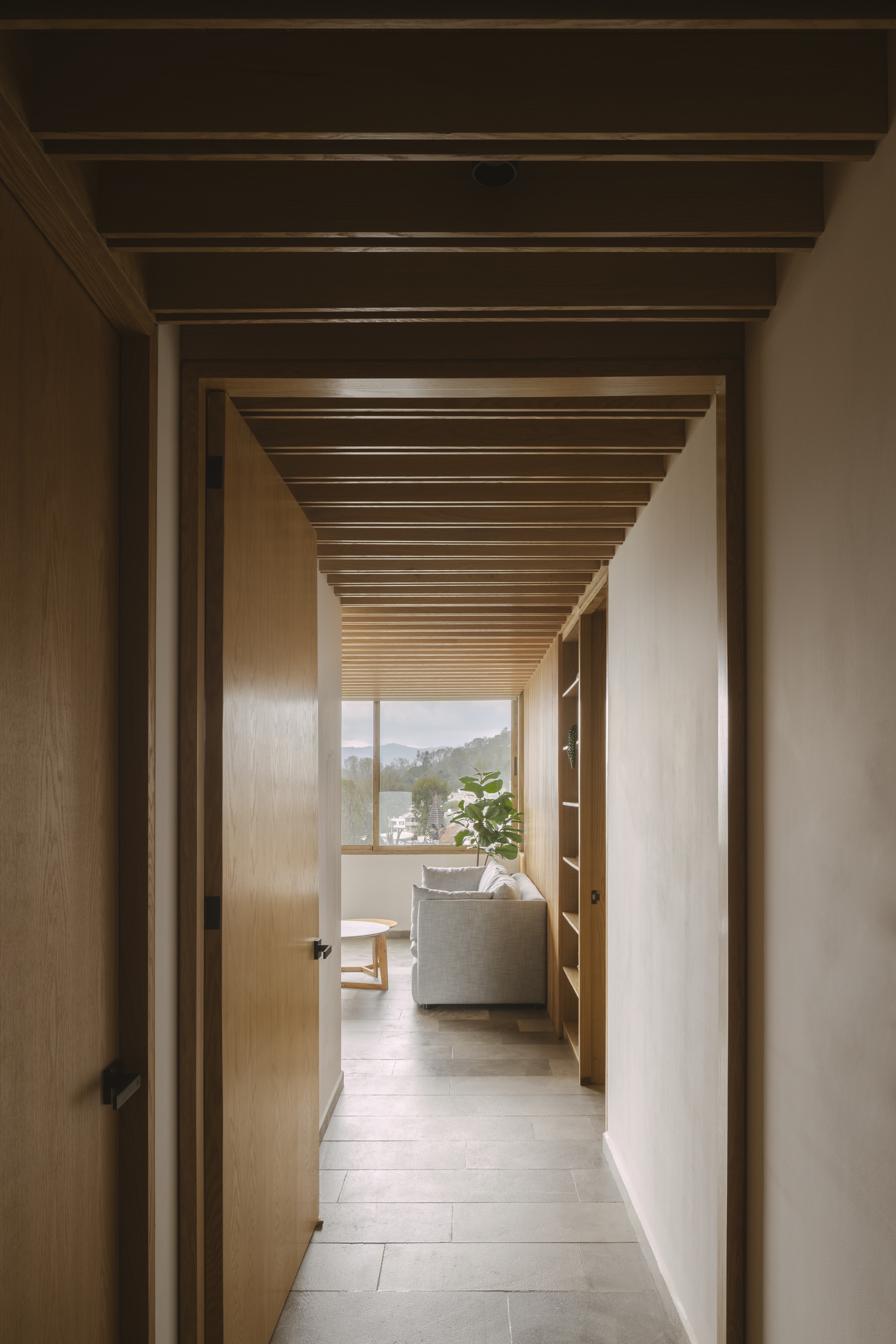 Timber-lined corridor with exposed ceiling beams leading to a grey sofa and a window onto green hills
