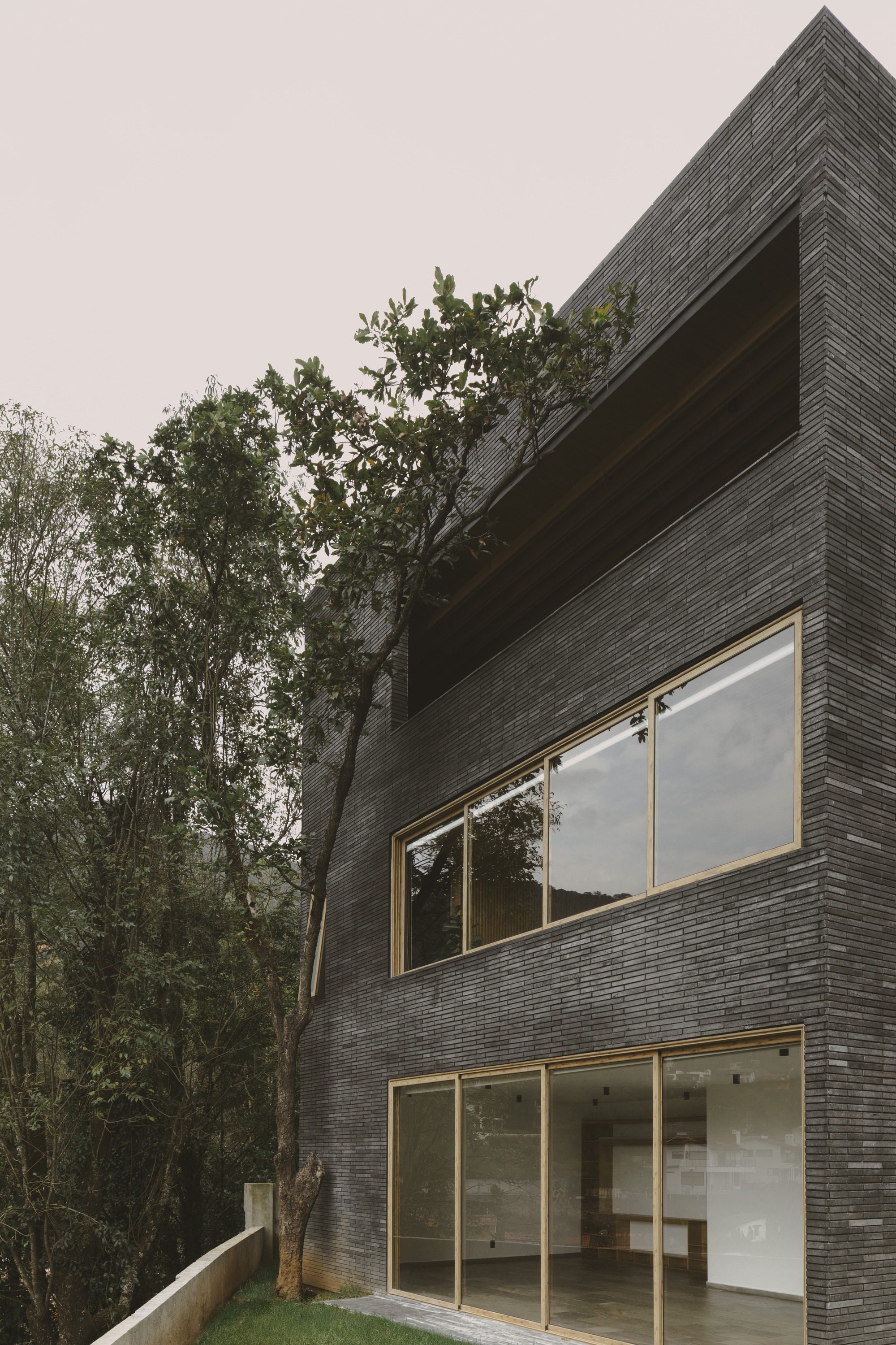 Dark brick facade with timber-framed glazing and a deep recessed opening, partly screened by a leaning tree under an overcast sky