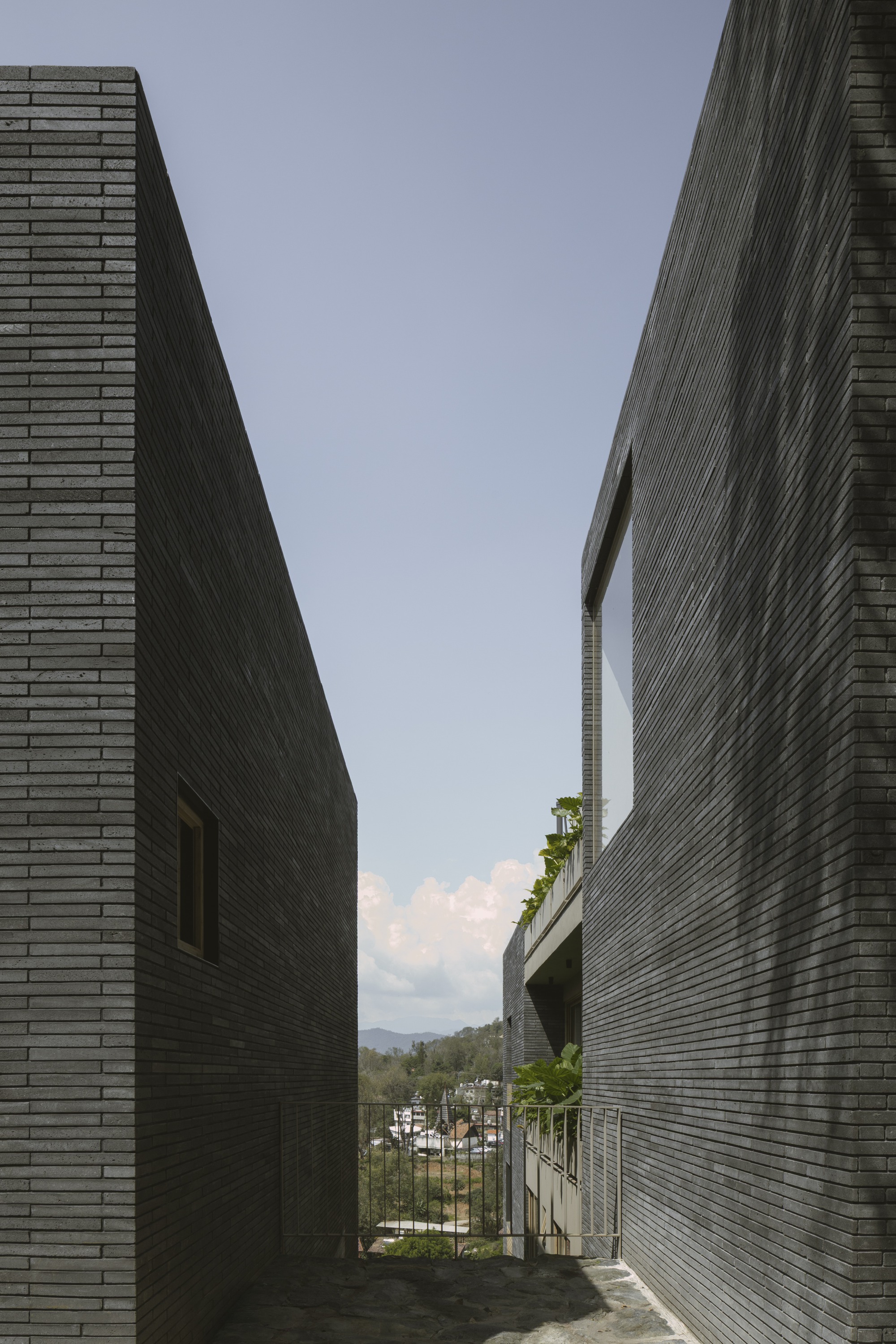 Narrow gap between two dark brick volumes framing a distant hillside, with planting spilling over a balcony in afternoon sun