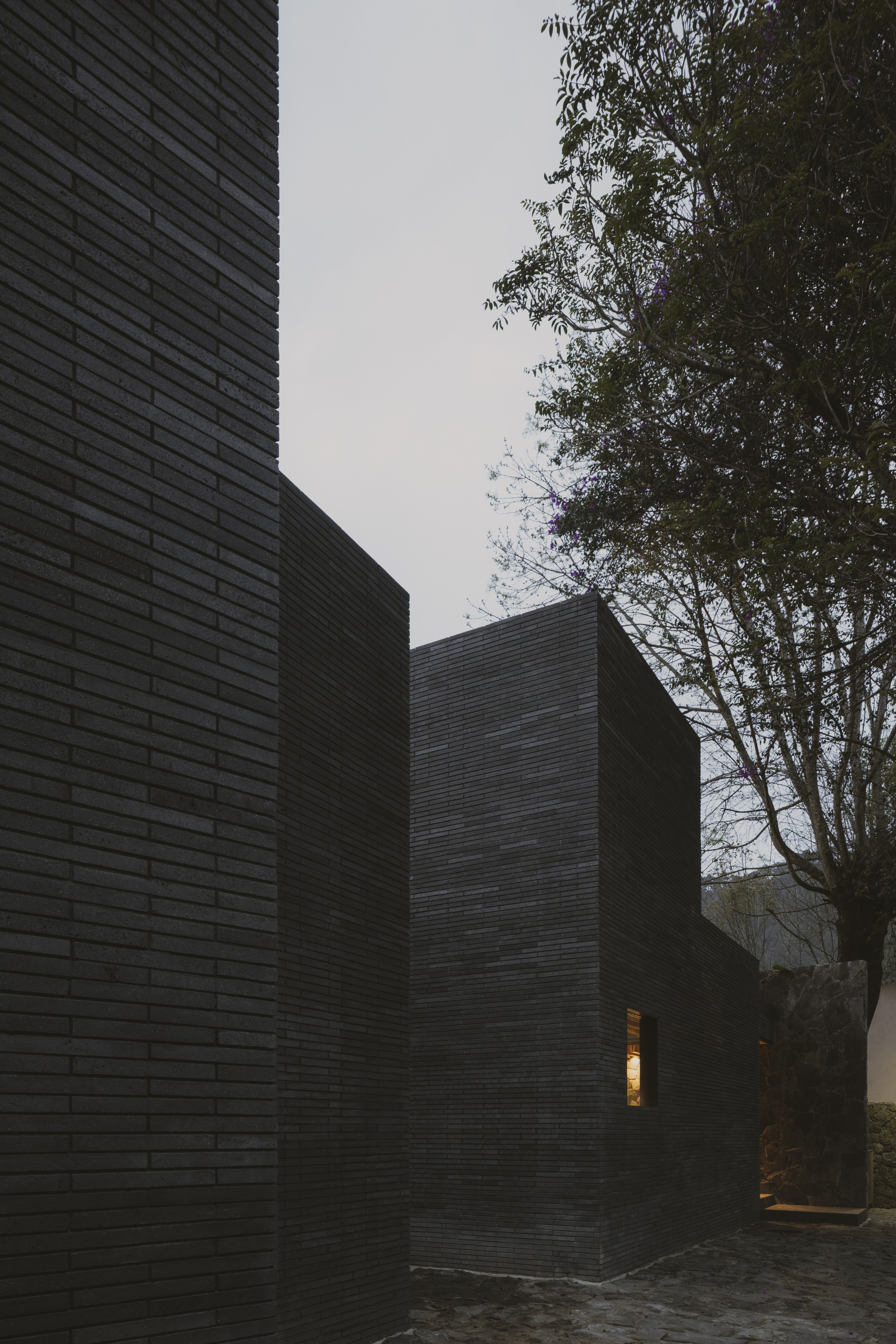 Stepped dark brick volumes at dusk with one small lit window and a jacaranda tree leaning over the cobbled court