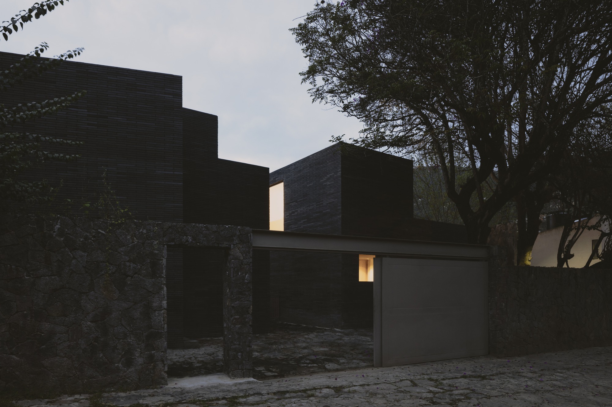 Street view of a stone gatepost and flat garage door before stacked dark brick volumes, with lit windows at dusk