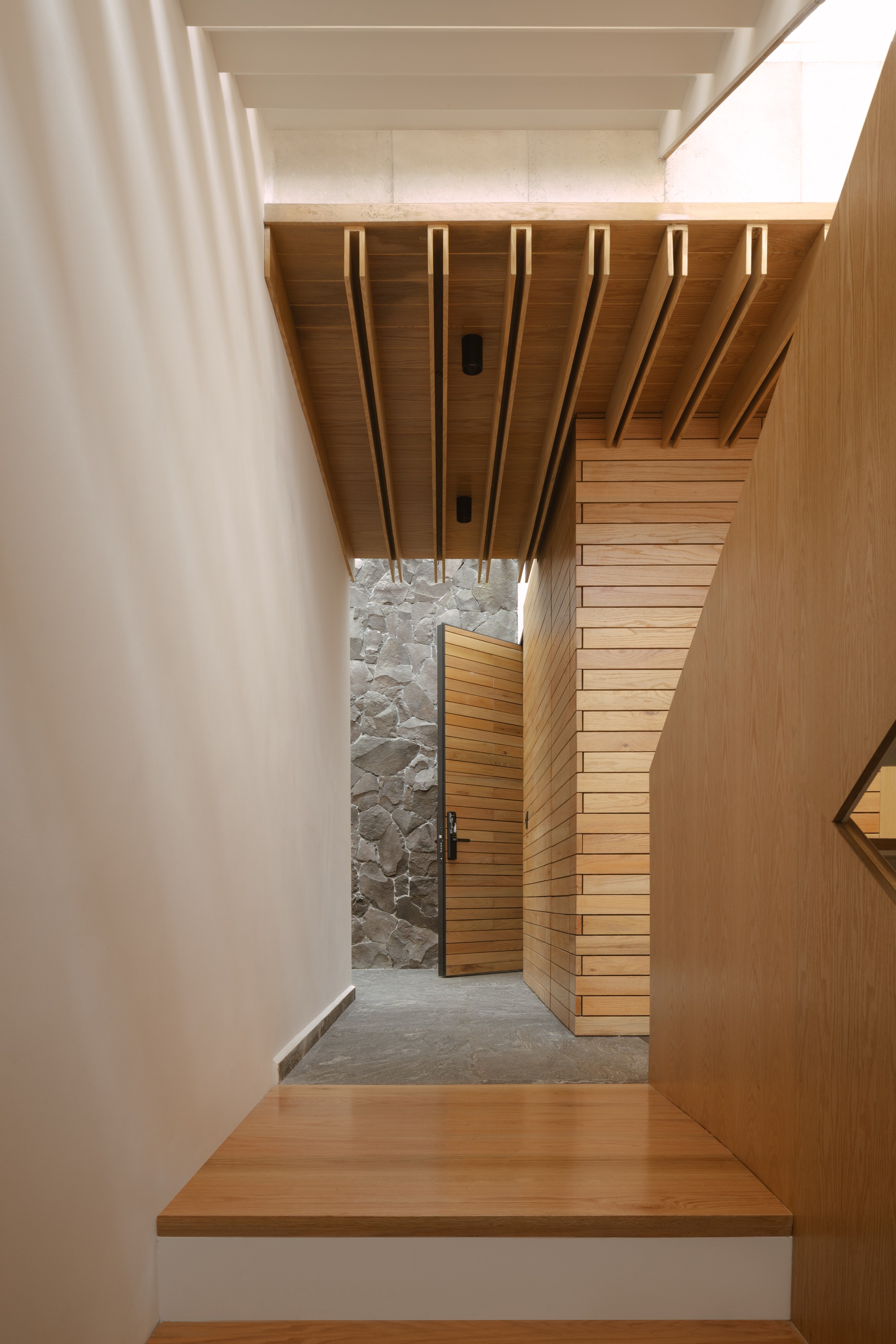 Timber-lined passage with exposed ceiling joists leading past a wood-slatted door to a rough stone wall, lit by a skylight