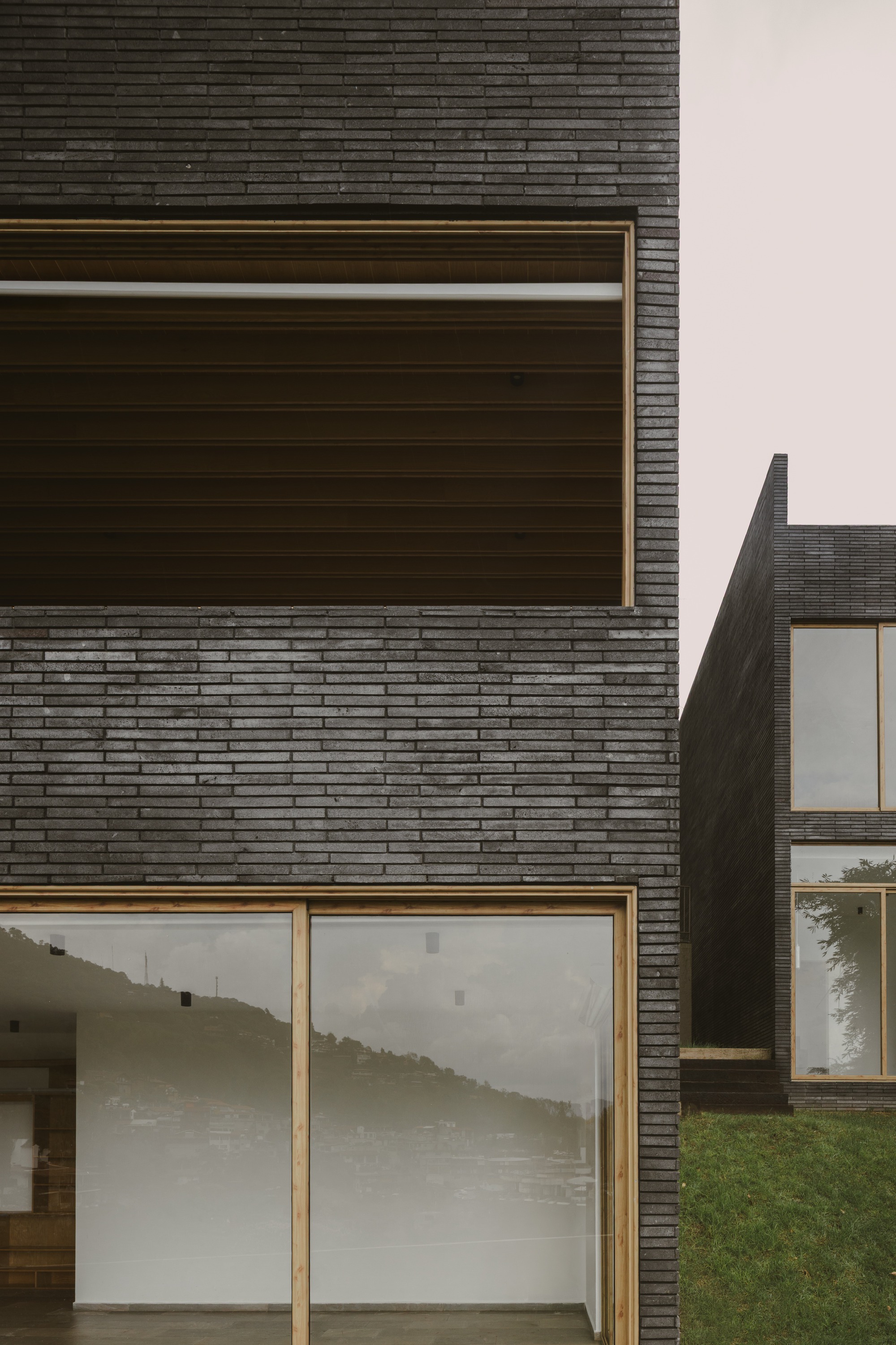 Close-up of dark long-format brick wall with timber-framed openings, hillside reflected in the glazing beneath a pale grey sky