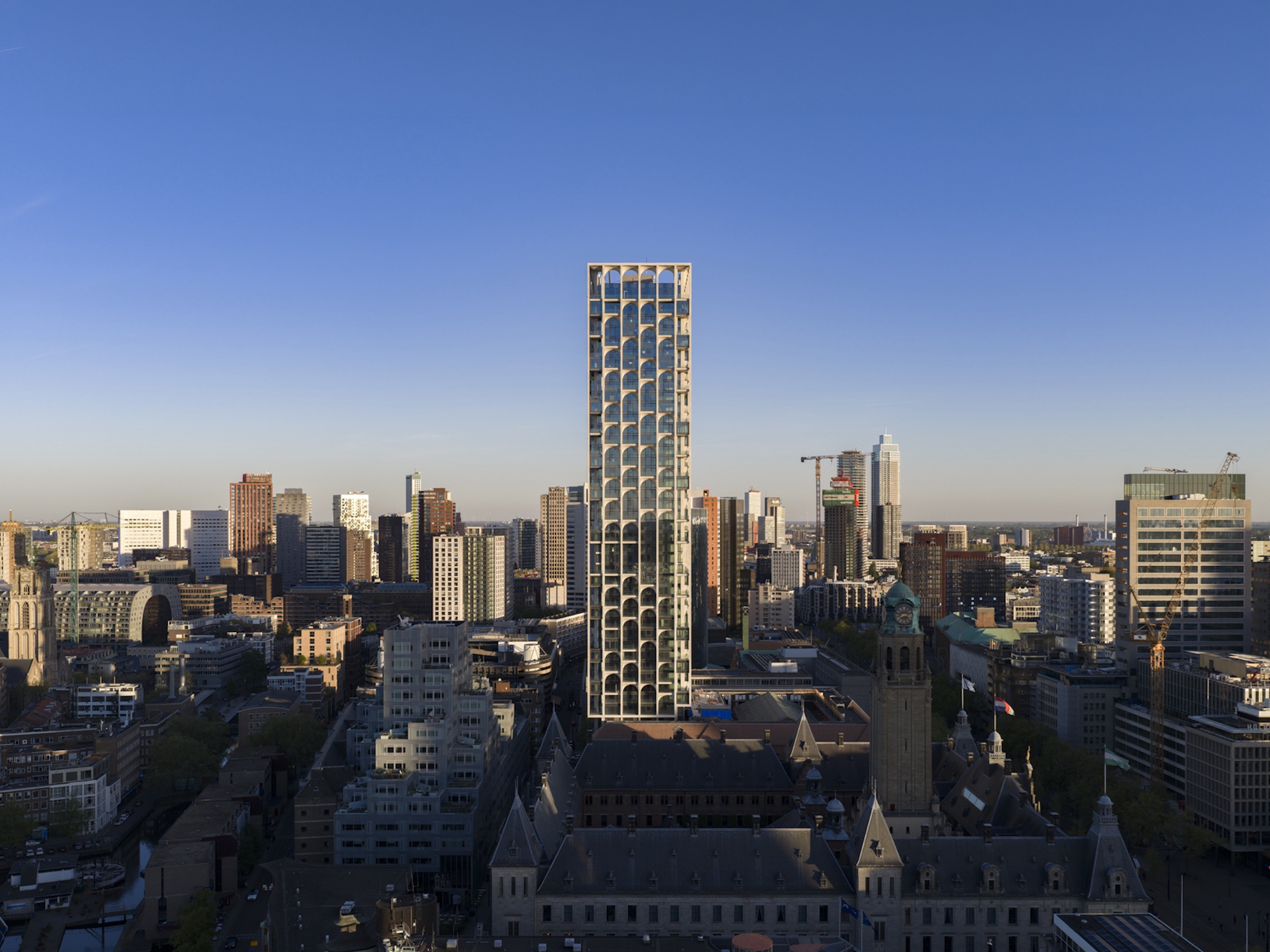 Tall tower with an arched white grid facade rising above a low city skyline and a historic civic building at dusk