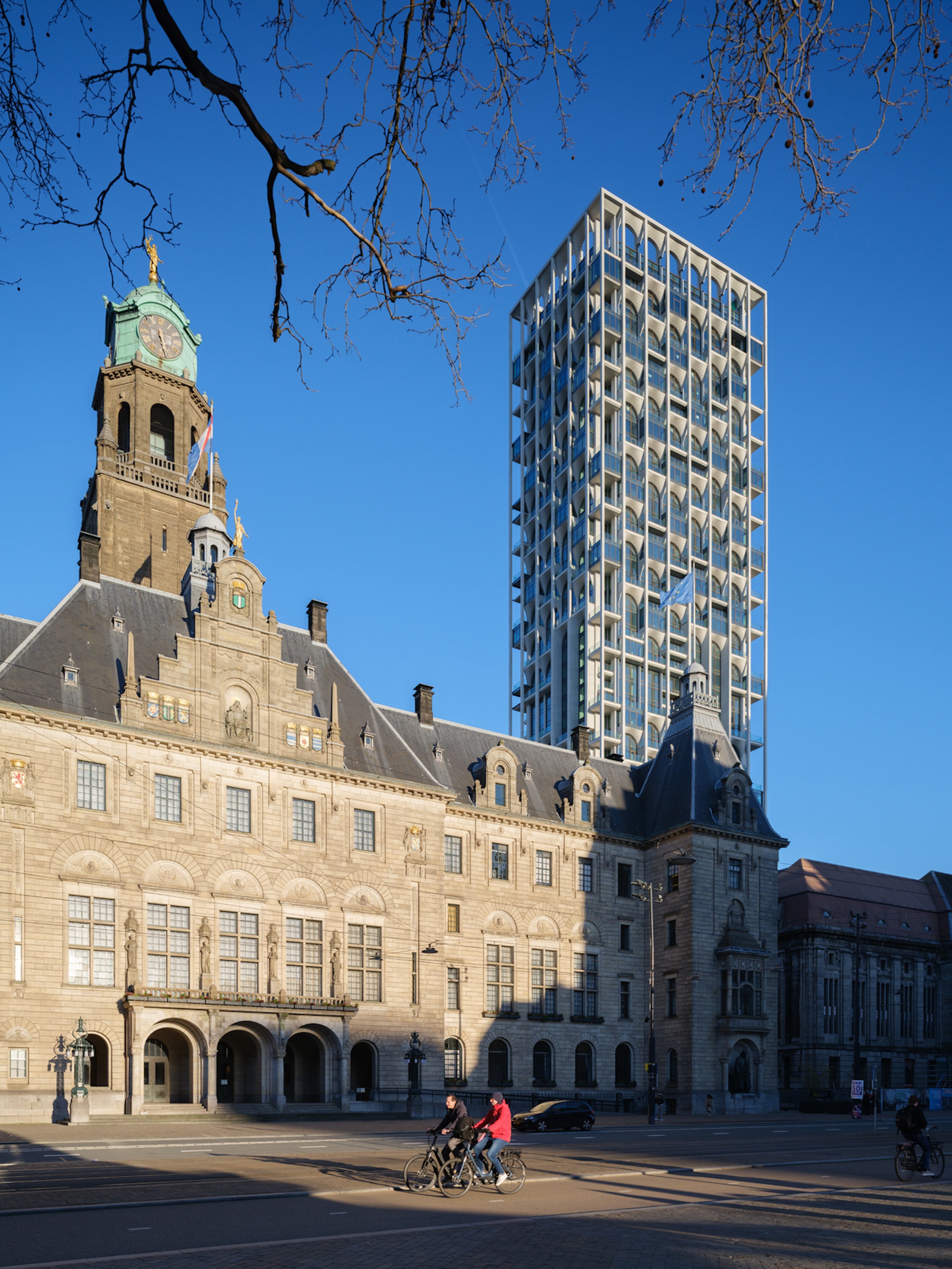 Glazed tower with projecting balconies rising behind a stone town hall with clock tower, two cyclists passing in low sun