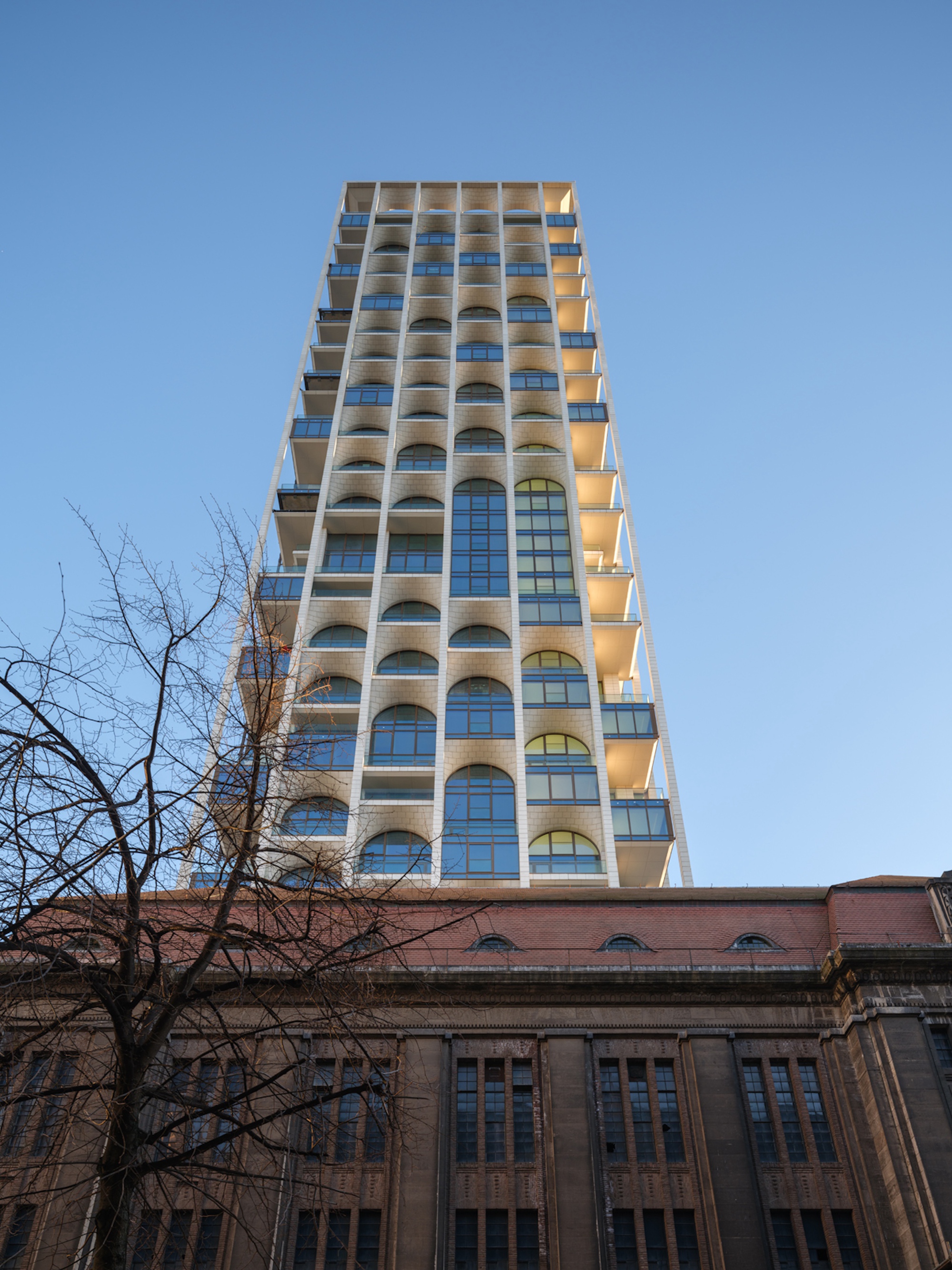 Upward view of the tower's arched windows, pale brick surfaces and cantilevered balconies above a brown stone roofline, with a bare tree in the foreground