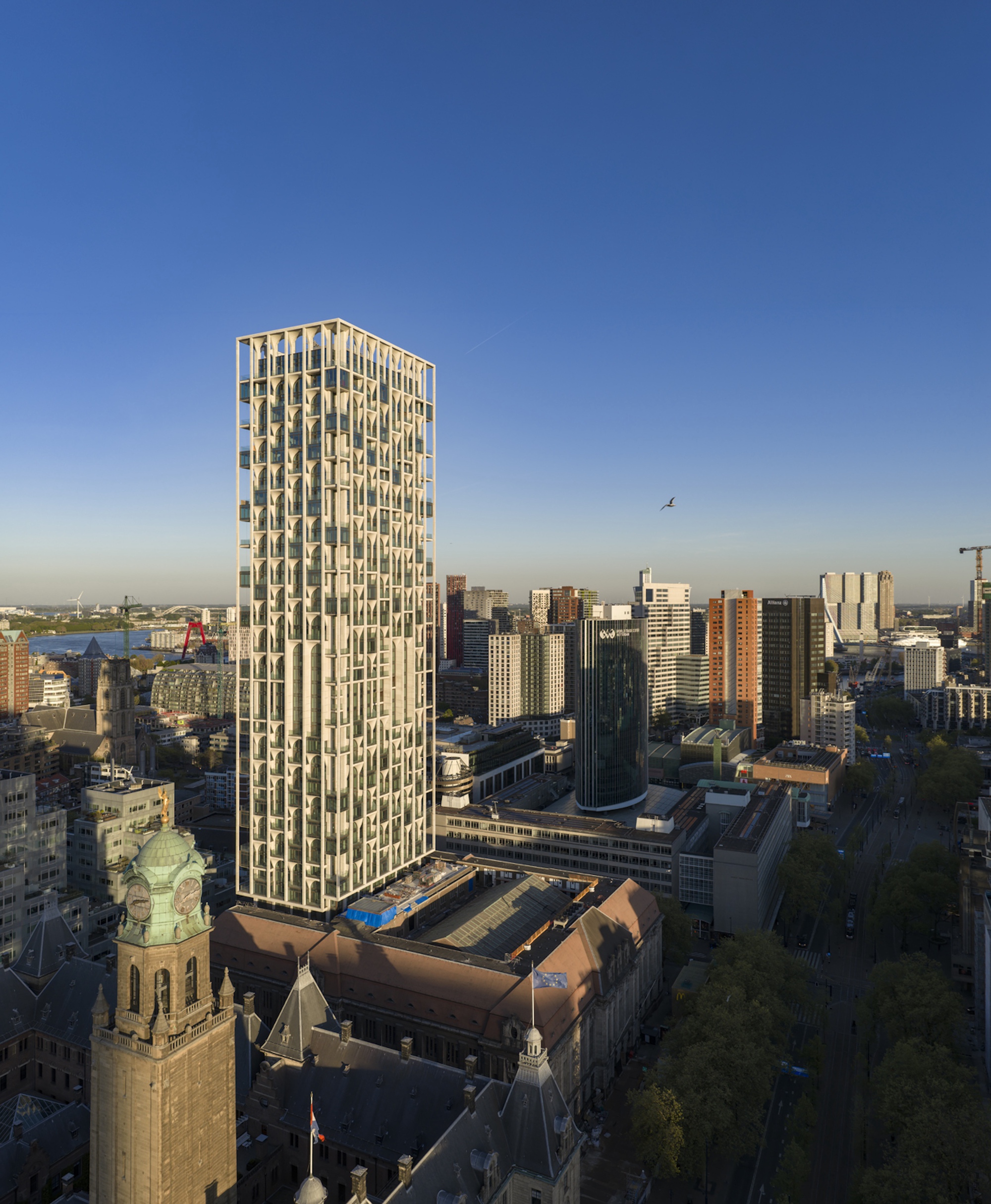 Aerial view of the gridded tower beside a green-domed clock tower and steep slate roofs in warm evening light