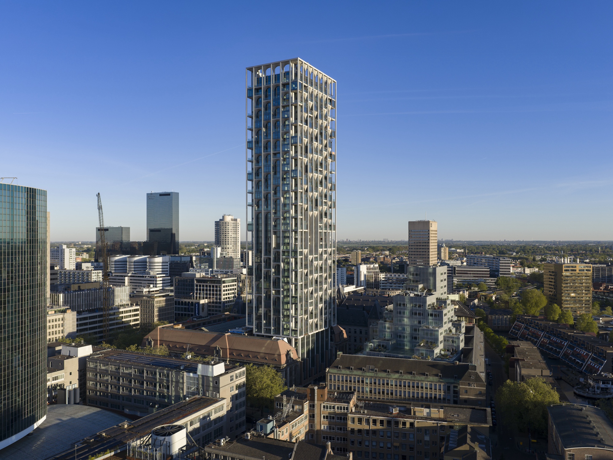 Slender tower with a white frame and recessed balconies standing above a dense neighbourhood of mid-rise blocks and treetops under a clear sky