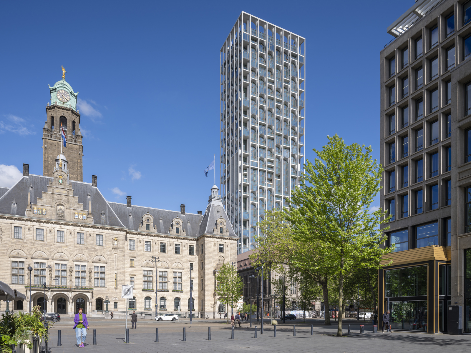 Tower with an arched white frame rising behind the town hall's clock tower, a leafy spring tree and a pedestrian crossing the square