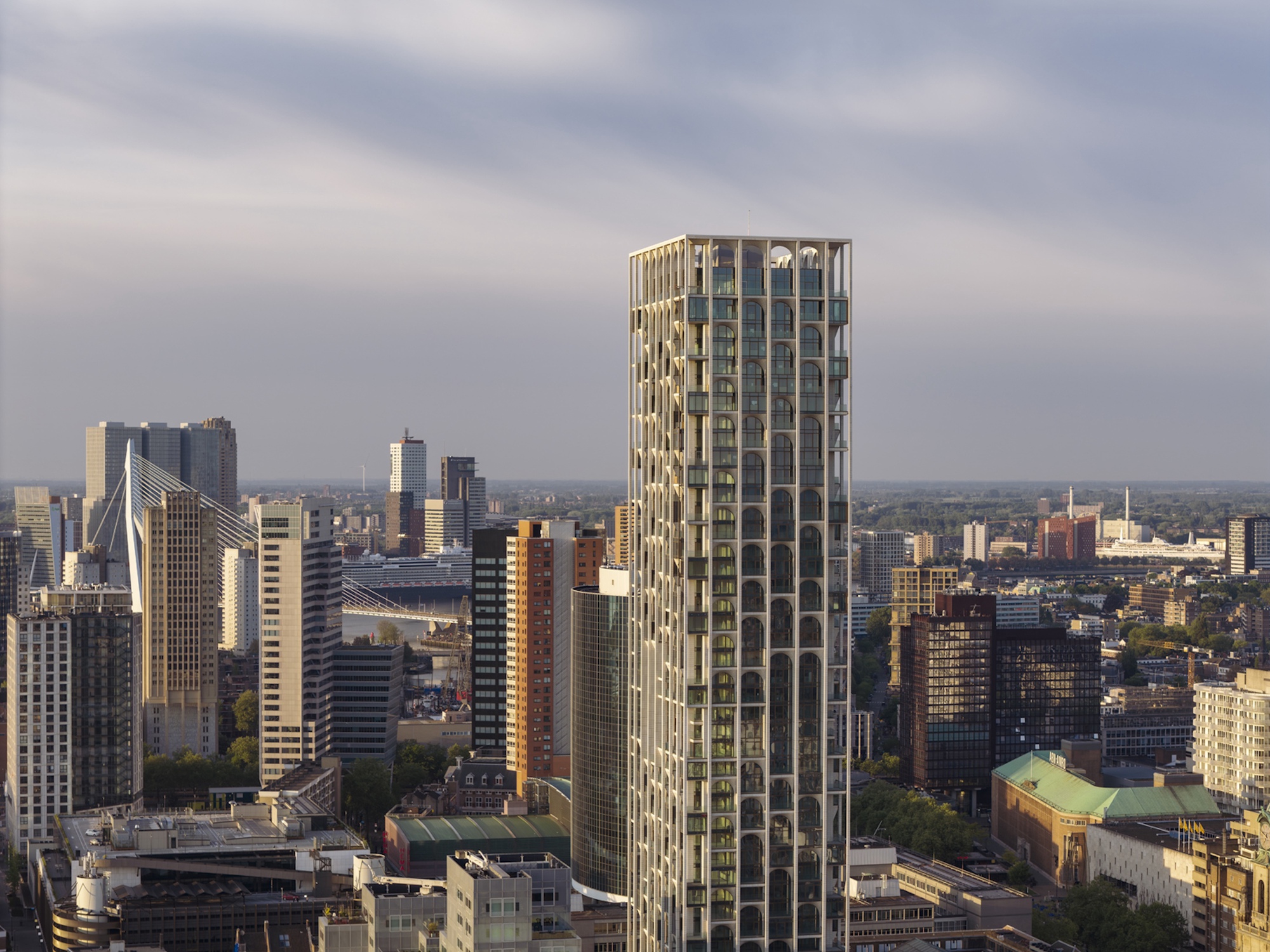 Aerial view of the arched-window tower among city blocks, with a cable-stayed bridge in the distance under heavy grey cloud