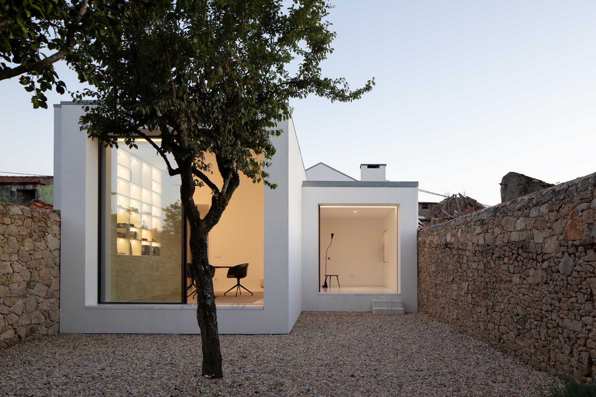 White box volumes with large glazed openings glowing at dusk behind a gnarled tree and gravel courtyard framed by stone walls
