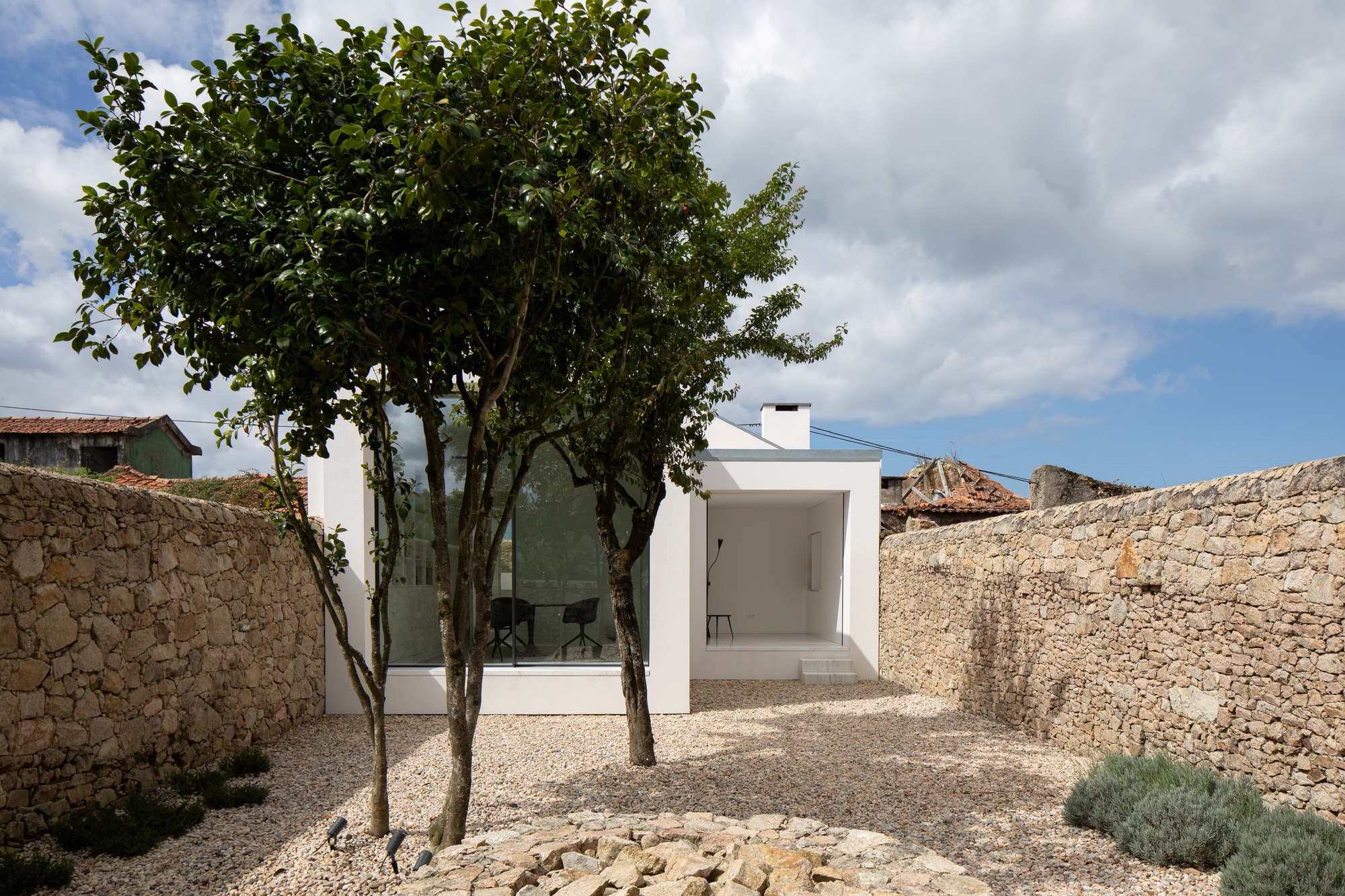 Gravel courtyard between two dry stone walls with a white pavilion seen through the branches of two trees under cloudy sky