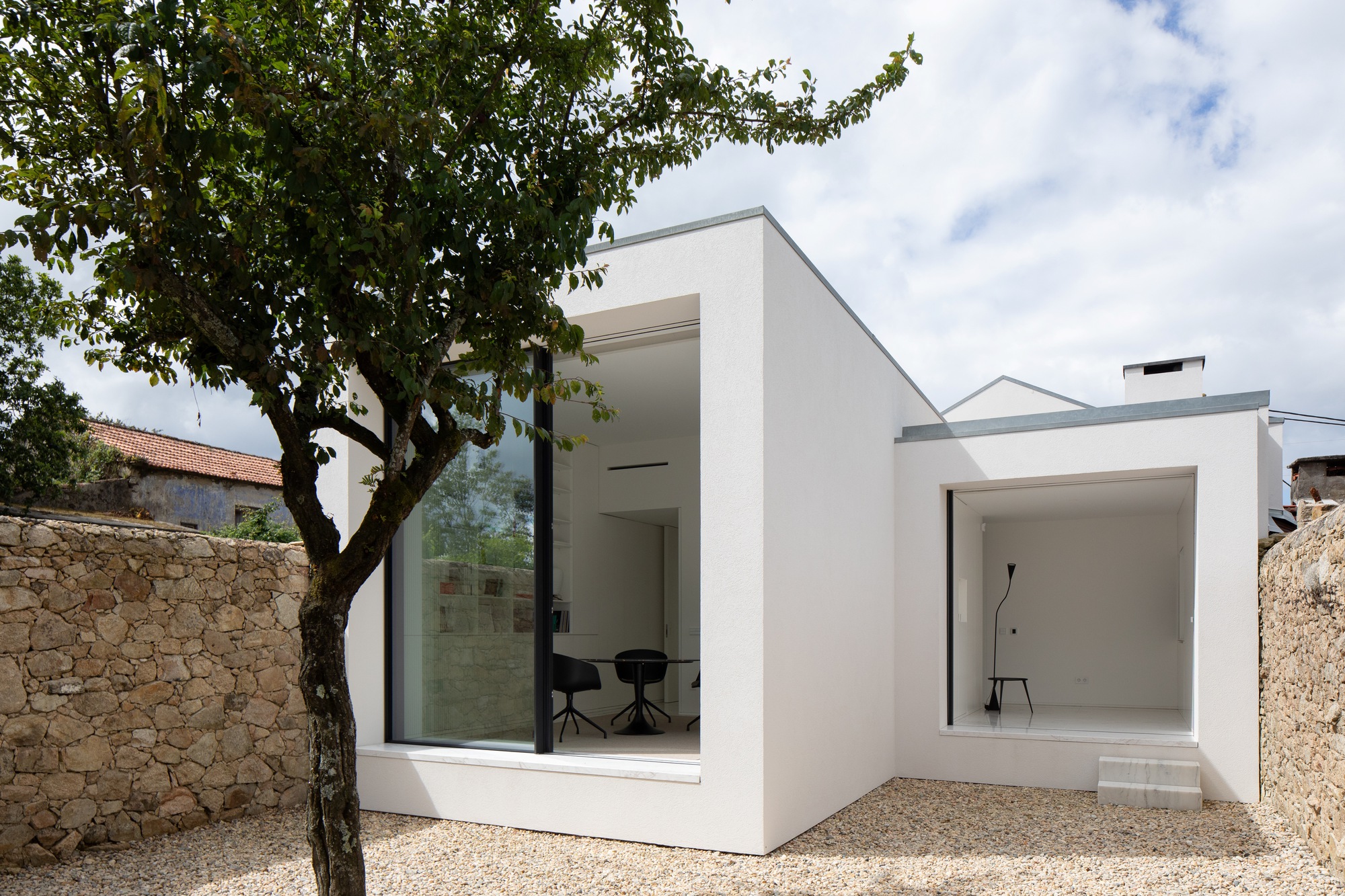 Corner view of two white rendered volumes with deep window reveals and a marble step, beside a tree trunk in daylight