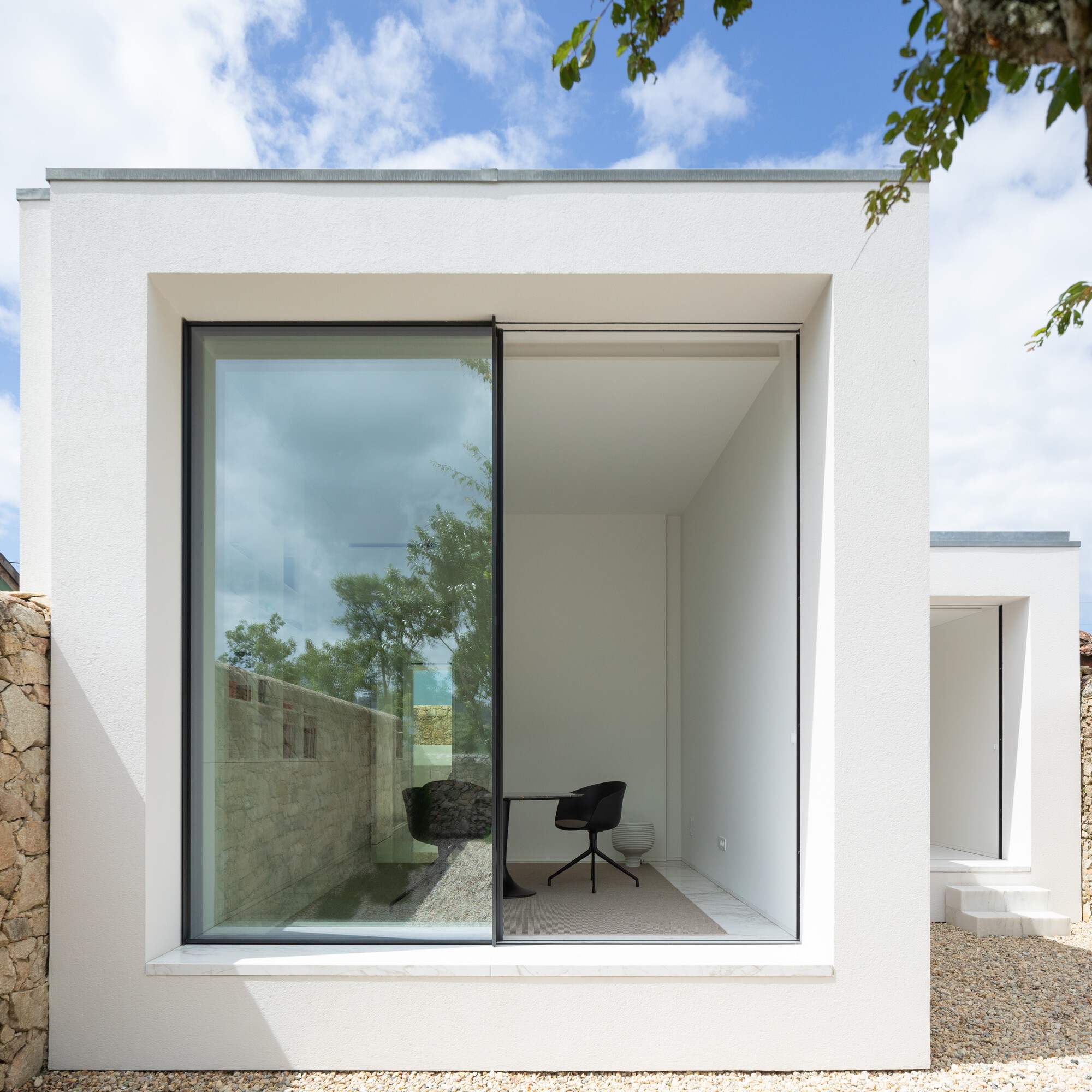 Frontal view of the white volume with a large sliding glass pane reflecting a stone wall and trees, black chair inside
